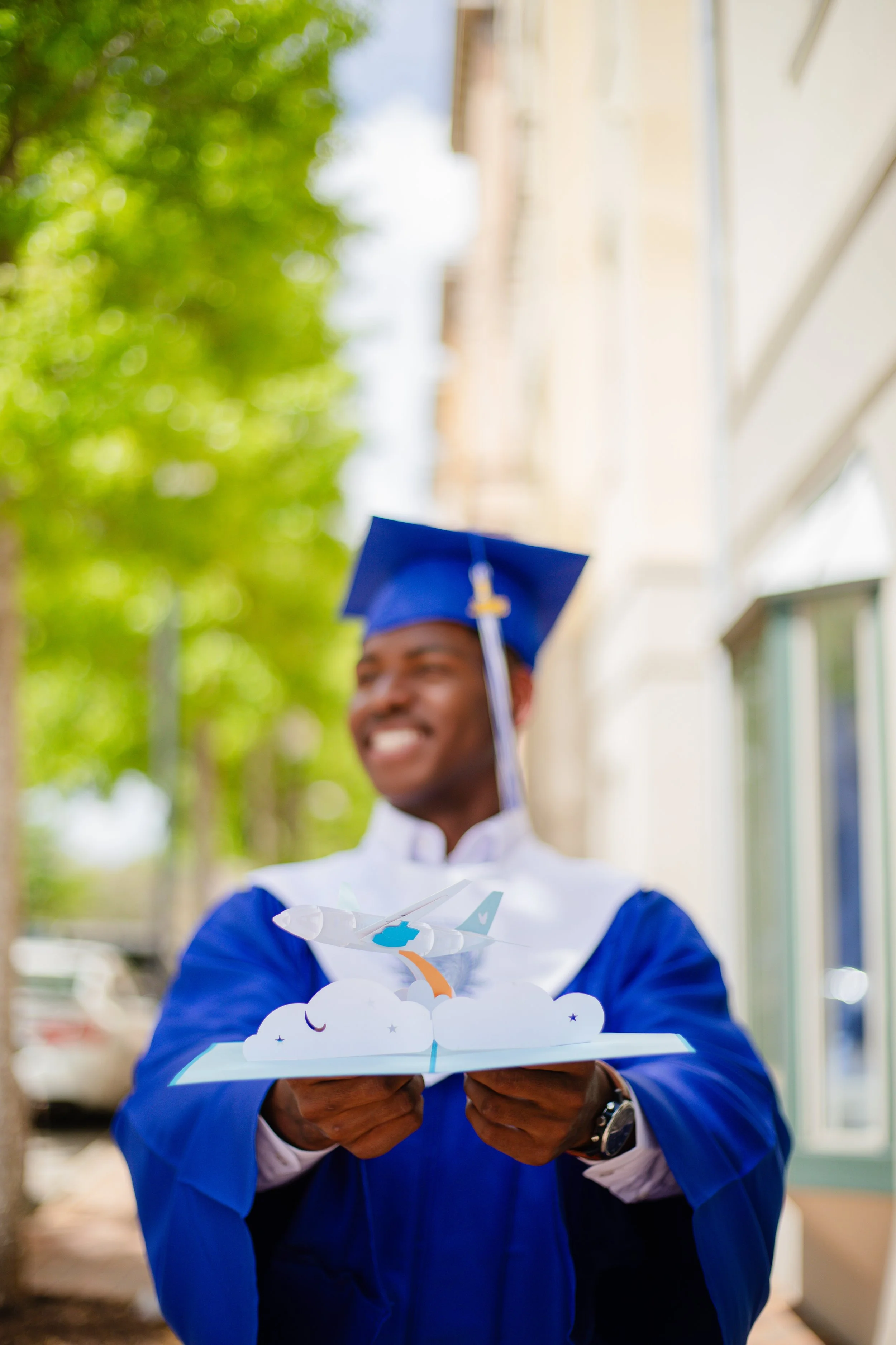 Graduate in blue cap and gown holding a pop-up paper airplane card outdoors.
