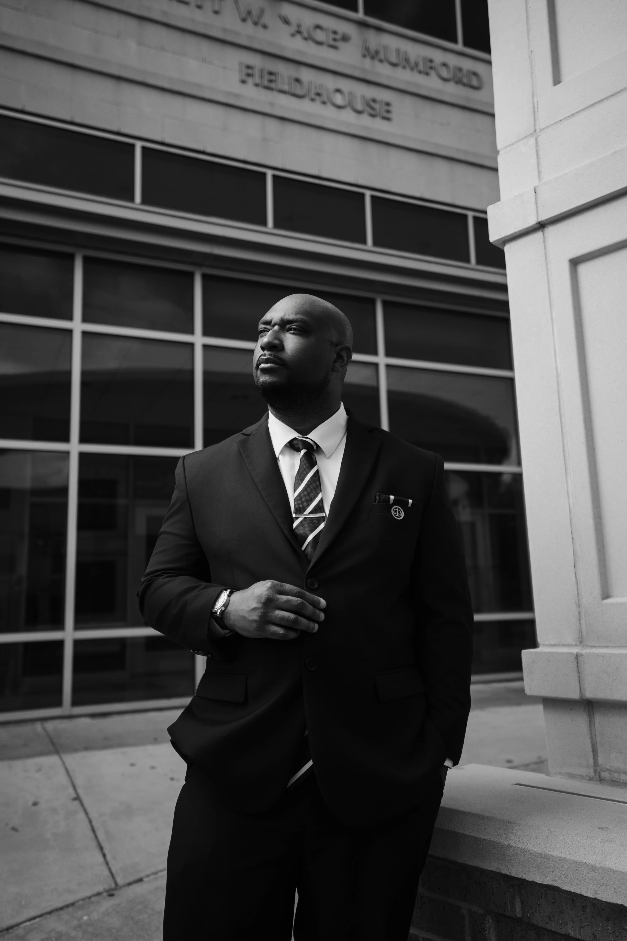 A black-and-white photo of a man in a business suit and tie, standing outdoors in front of a modern building with large windows and signage. He has a serious expression and is looking slightly upward and to the right.