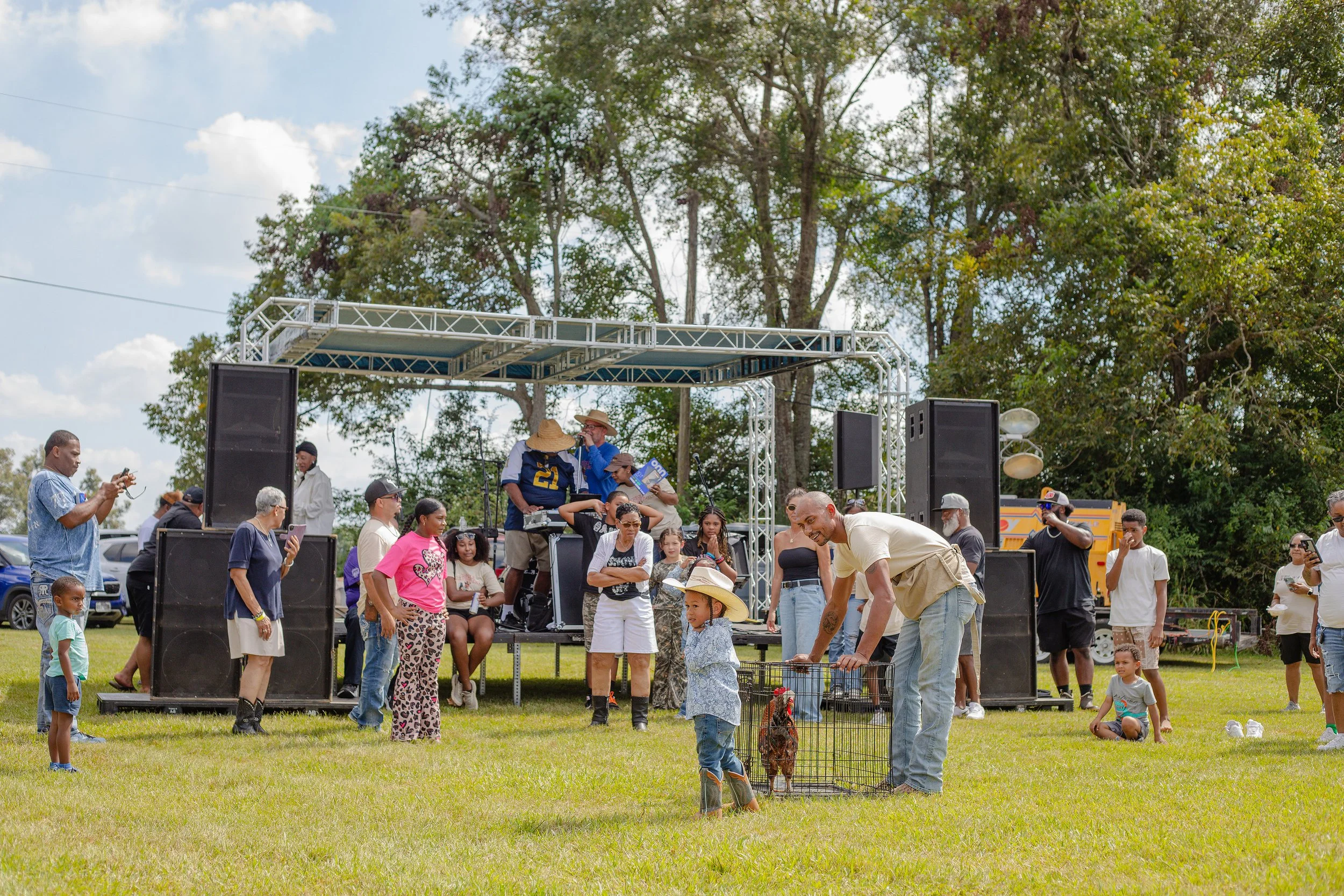 A diverse group of people gather outdoors on a grassy field, watching a small stage where musicians perform. A man is helping a child with a chicken in a cage. Some people are taking photos, and children are sitting on the grass. Trees and blue sky a
