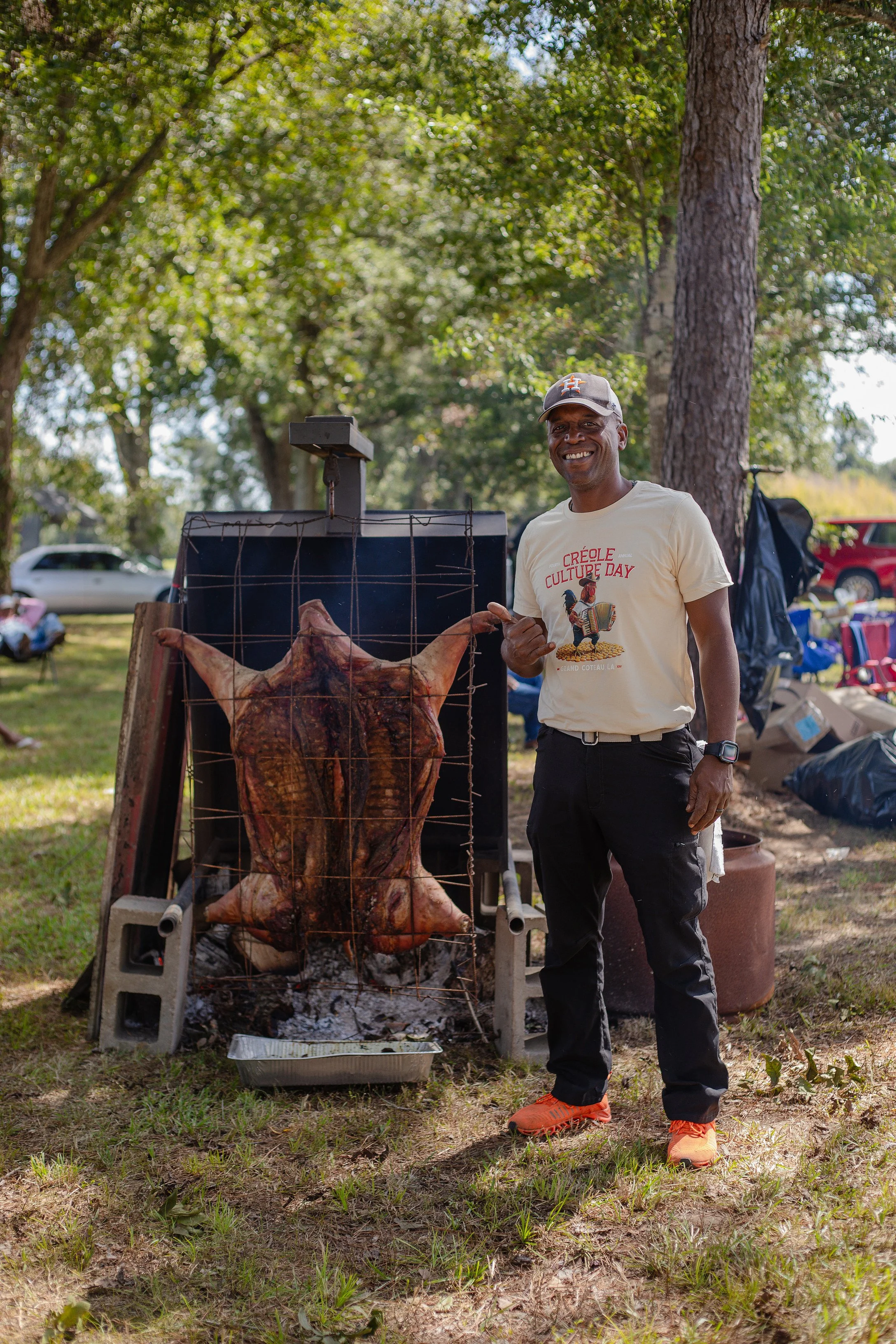 A man smiling outdoors in a park, standing next to a whole roasted pig on a grill.