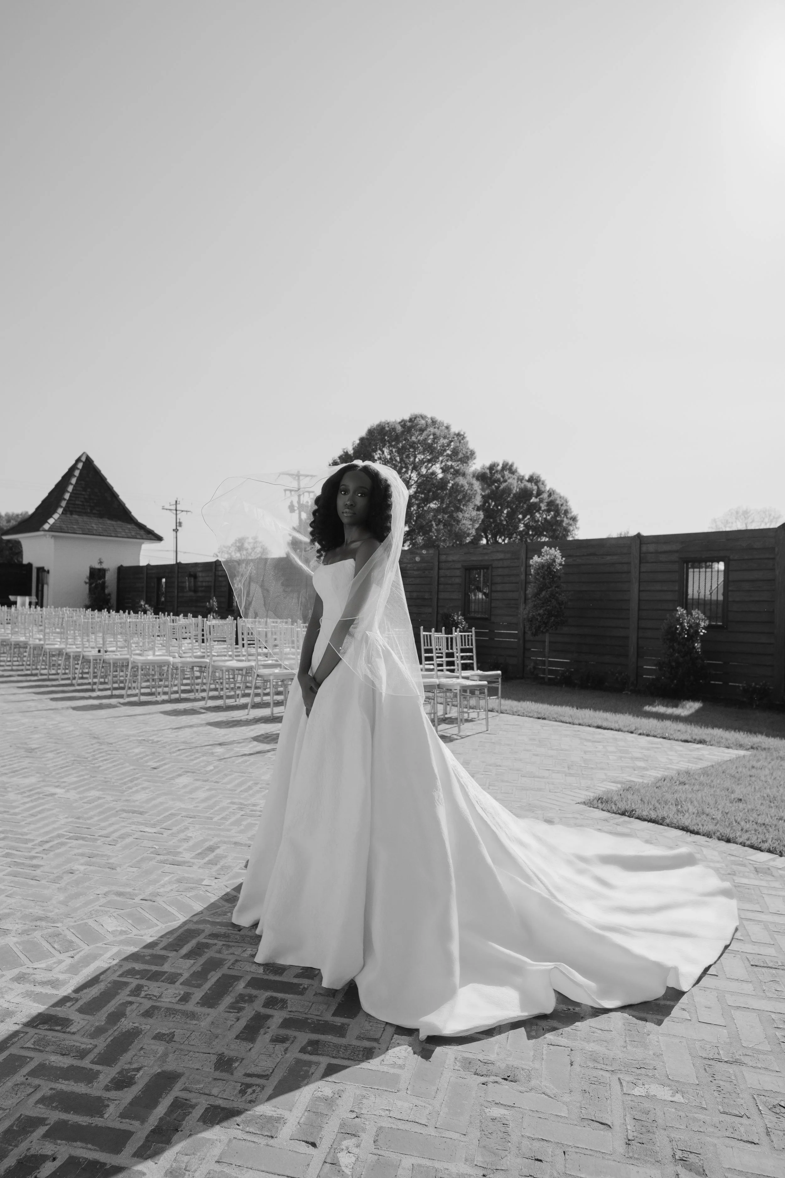 A bride in a strapless wedding dress with a long train and veil standing outdoors on a paved area, with chairs and a wooden fence in the background, captured in black and white.
