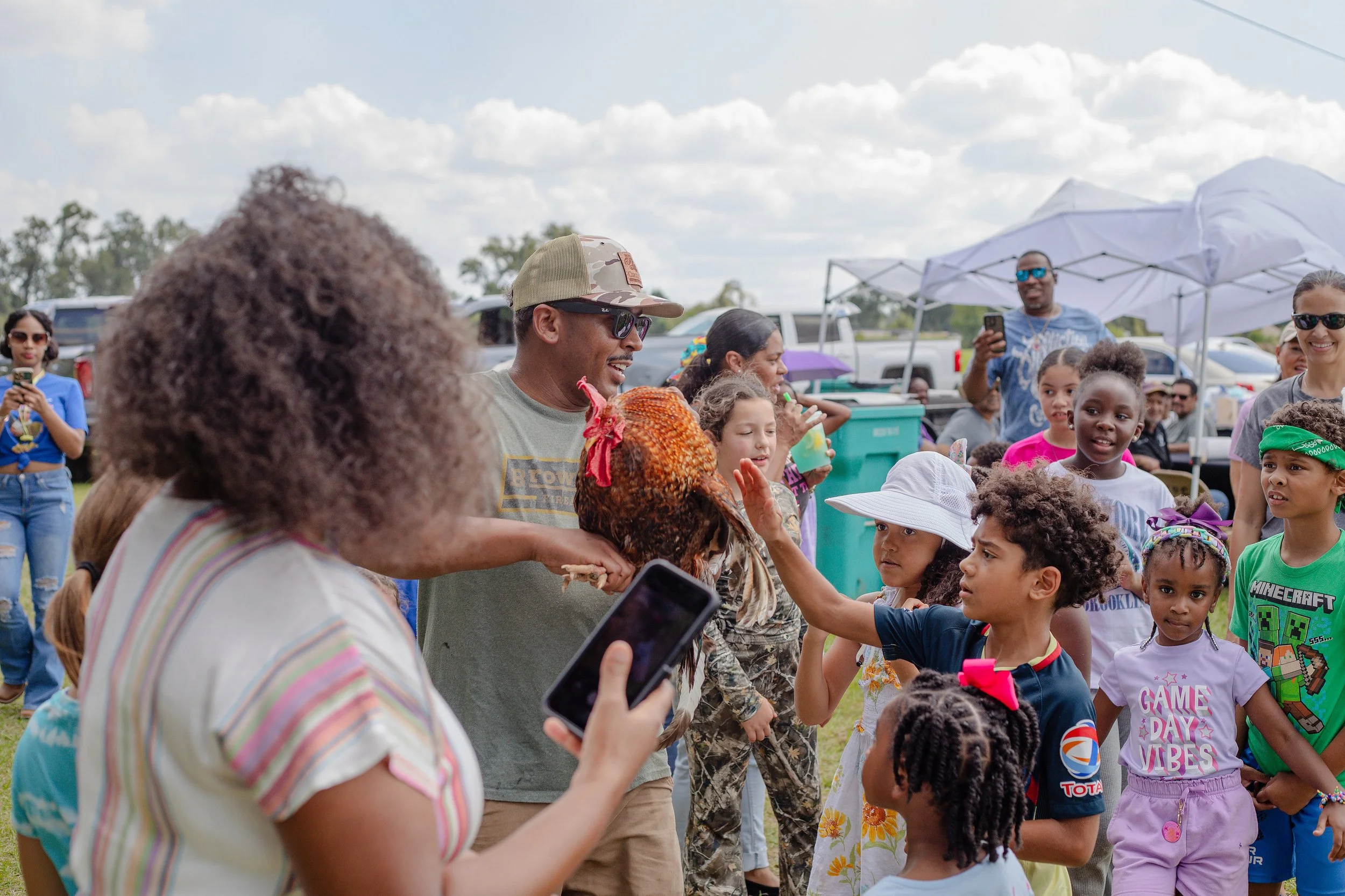 Group of children and adults gathered outdoors at a community event, interacting with a person holding a brown chicken, with tents and parked cars in the background.