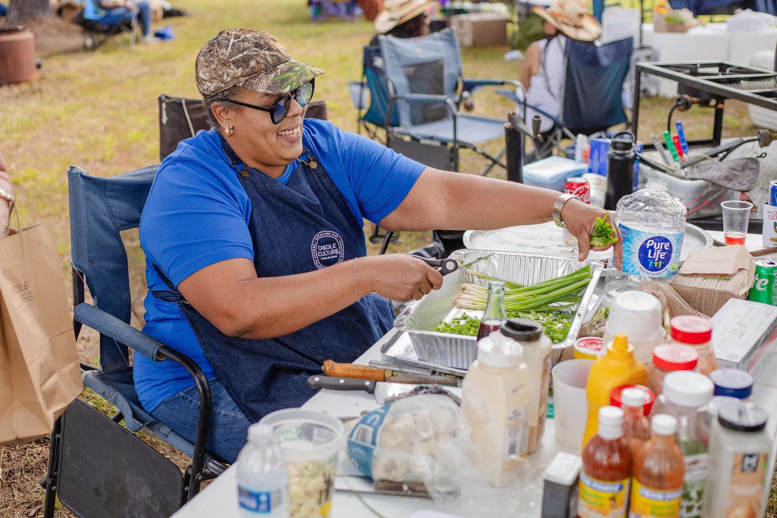 Woman cooking at an outdoor event, chopping green onions with a smile, surrounded by food supplies and condiments on a table.