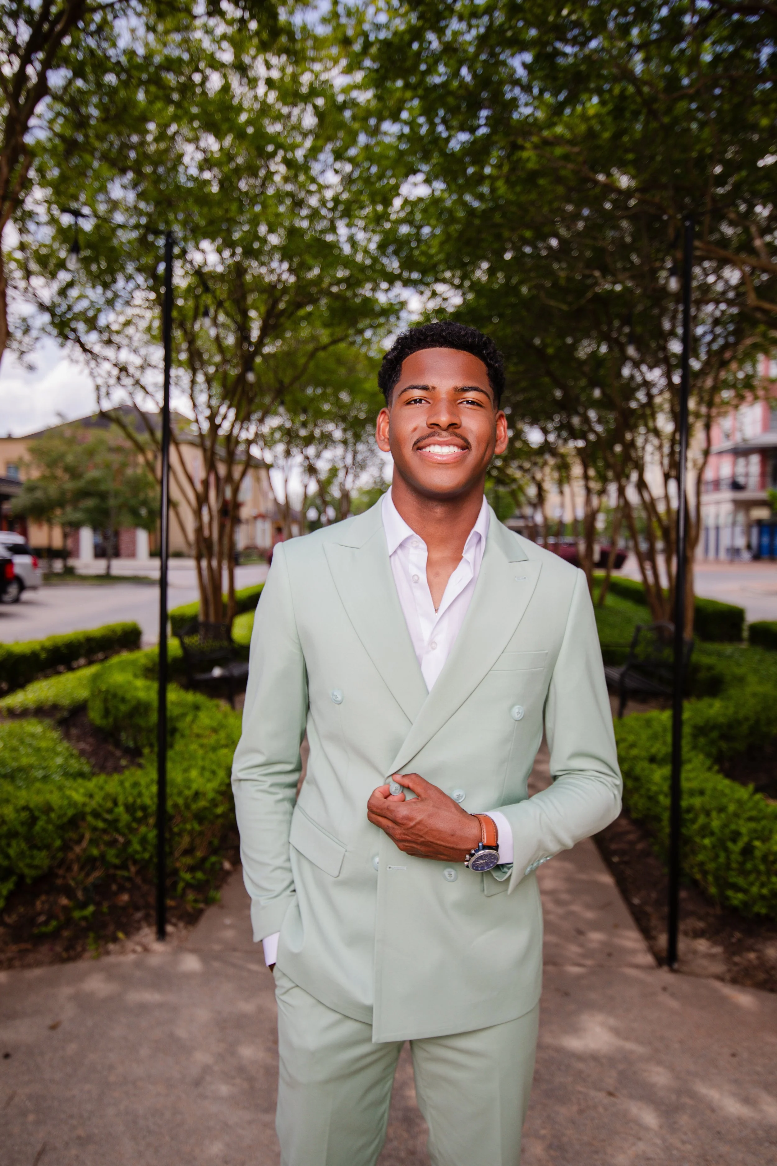 A young man in a light-colored suit standing outdoors in a park with green trees and a bench in the background.