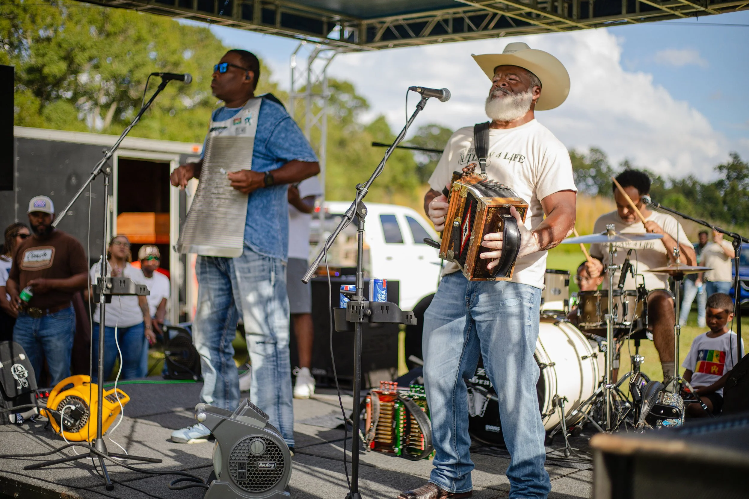 Musicians performing outdoors at a festival. One man with a beard wearing a cowboy hat plays an accordion, another man wearing sunglasses plays a washboard, and a drummer is in the background. Audience members watch the performance.