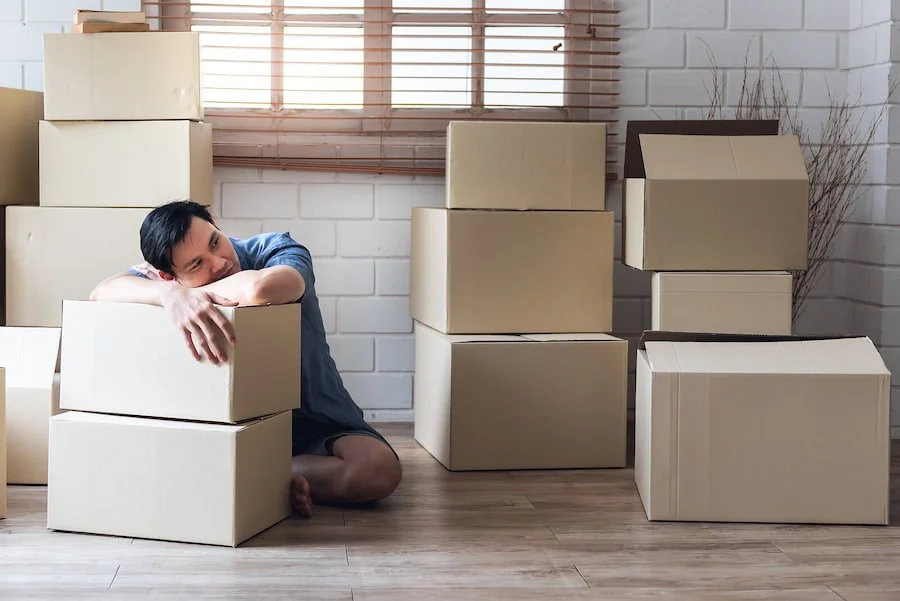 Image of a man sitting on the floor of an apartment with moving boxes around him. This image represents the life changes that people went through during the pandemic, which can be processed with a therapist in counseling for life transitions in San …