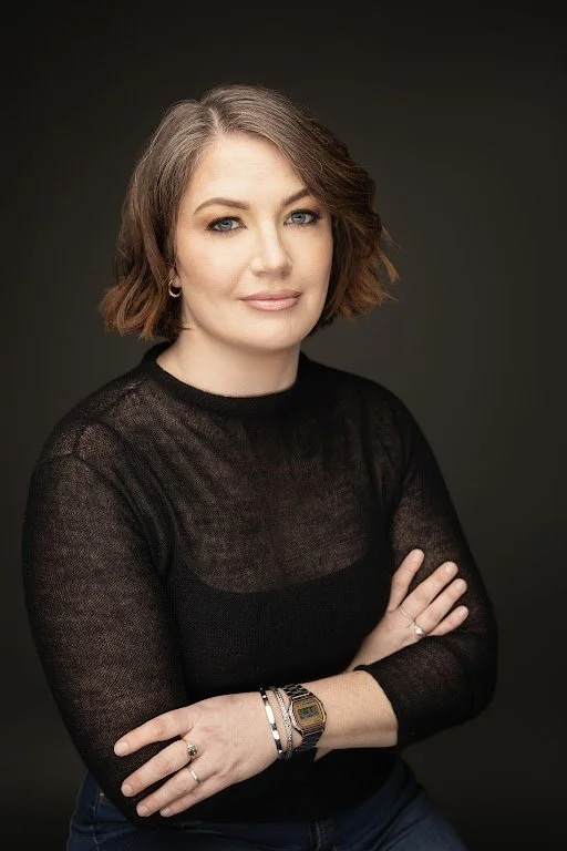 A woman with short, wavy, light brown hair and blue eyes, wearing a black sheer long-sleeving top and multiple bracelets, poses against a dark background.