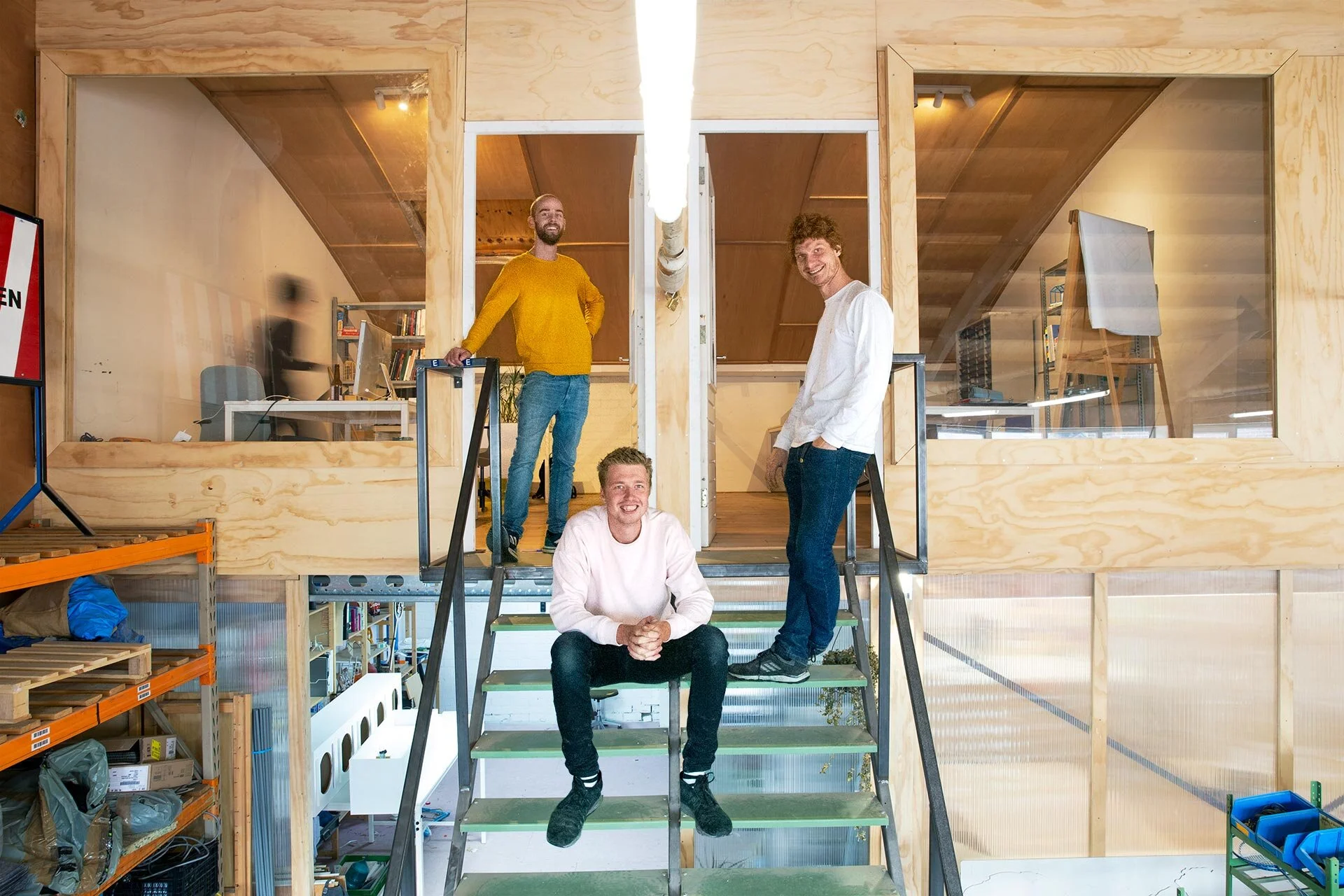 Vier mannen in een houten kantoorruimte met trappen, lachen en poseren voor de camera, twee binnen en twee buiten