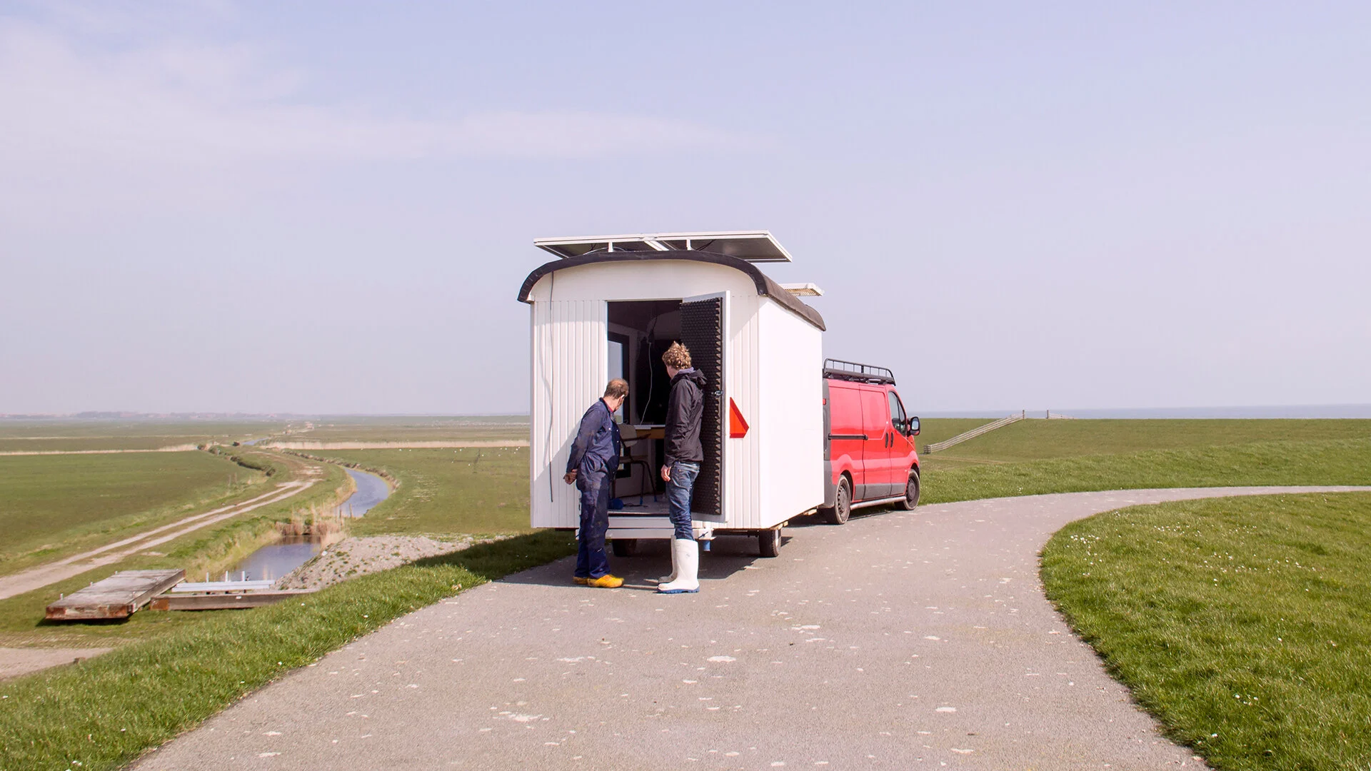 Twee mensen staan voor een kleine witte mobiele werkunit op wielen, gekoppeld aan een rode bus, op een landweg met groene velden en een kleine waterweg in de achtergrond.