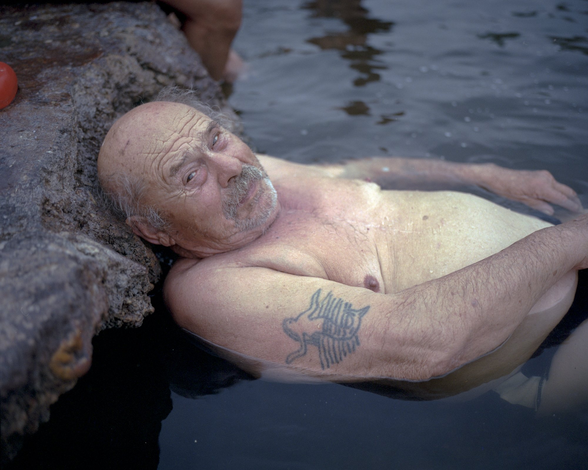  Bather with a mermaid tattoo. Törtel, Hungary, 2022   “I am near Törtel in the middle of nowhere. I sit in a healing thermal water spring, on the site of an abandoned oil well where water has burst up instead of black gold in the 60s, much to the de
