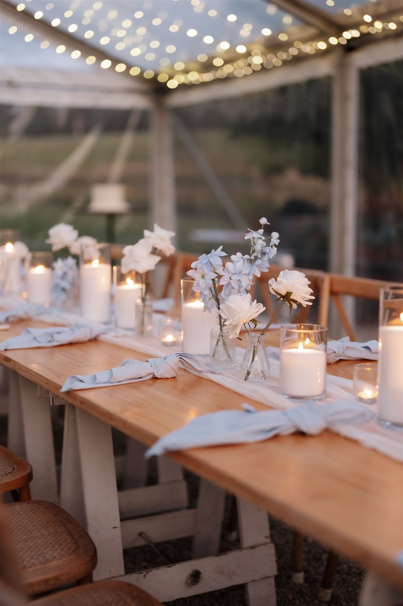 Table decorated with white flowers, candles, and napkins, set for an outdoor event under a canopy with string lights.