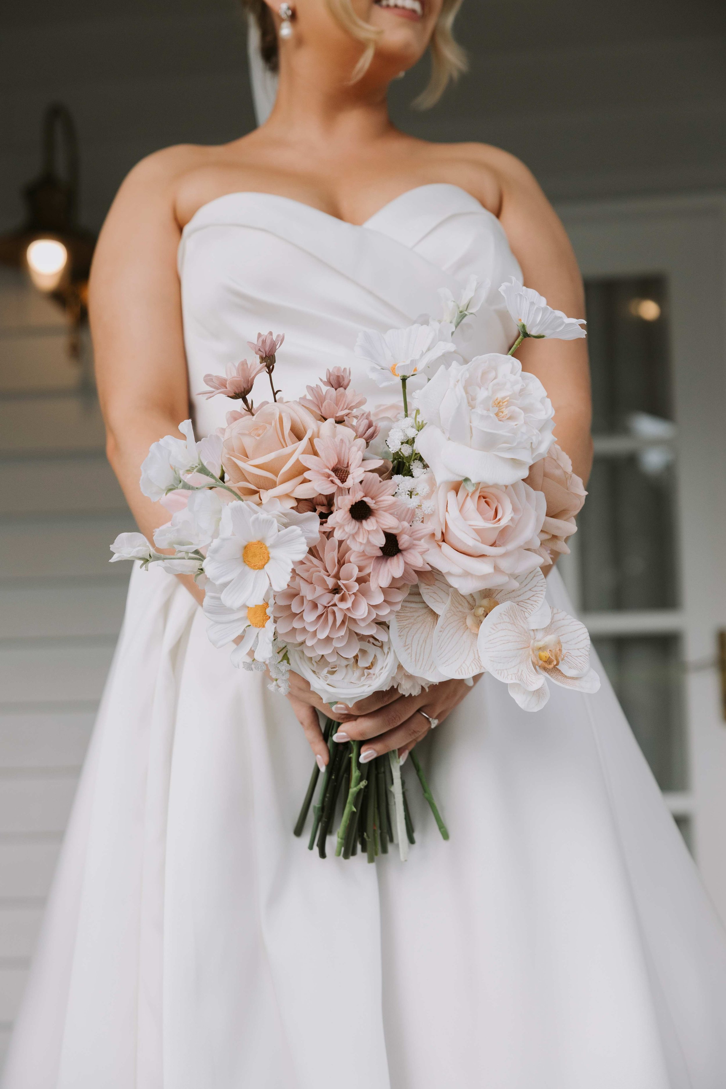 A bride in a white strapless wedding dress holding a bouquet of pink and white flower bouquet made out of artificial flowers. The bouquet has orchids, roses and cosmos.