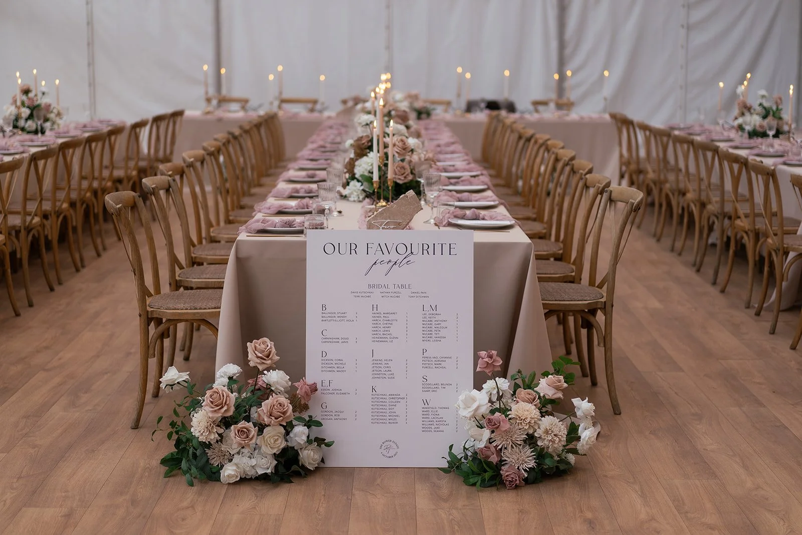 Wedding reception setup with long banquet table, latte, coffee and white flowers, candles, and seating options, with a seating chart sign at the front. Table end flowers at the base of the table.