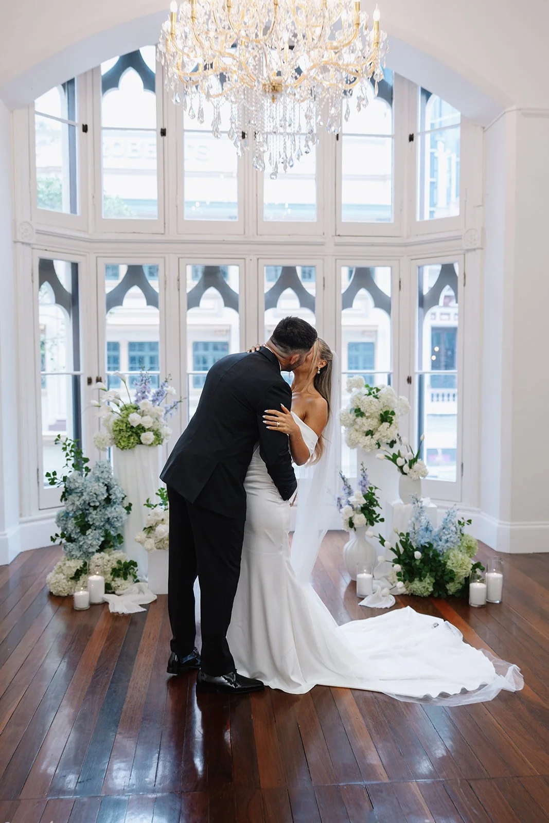 Bride and groom kissing during their wedding ceremony in an elegant room with large windows, faux floral arrangements on plinths and floor, candles, and a chandelier overhead.