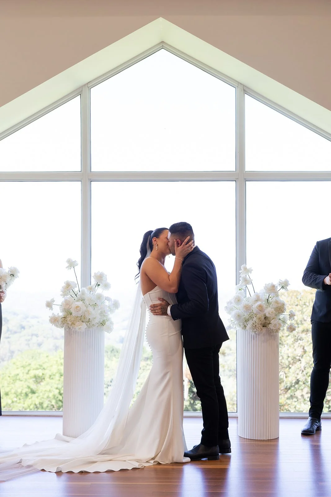 A bride and groom sharing a kiss during their wedding ceremony inside a modern chapel with large windows and white artificial floral arrangements.