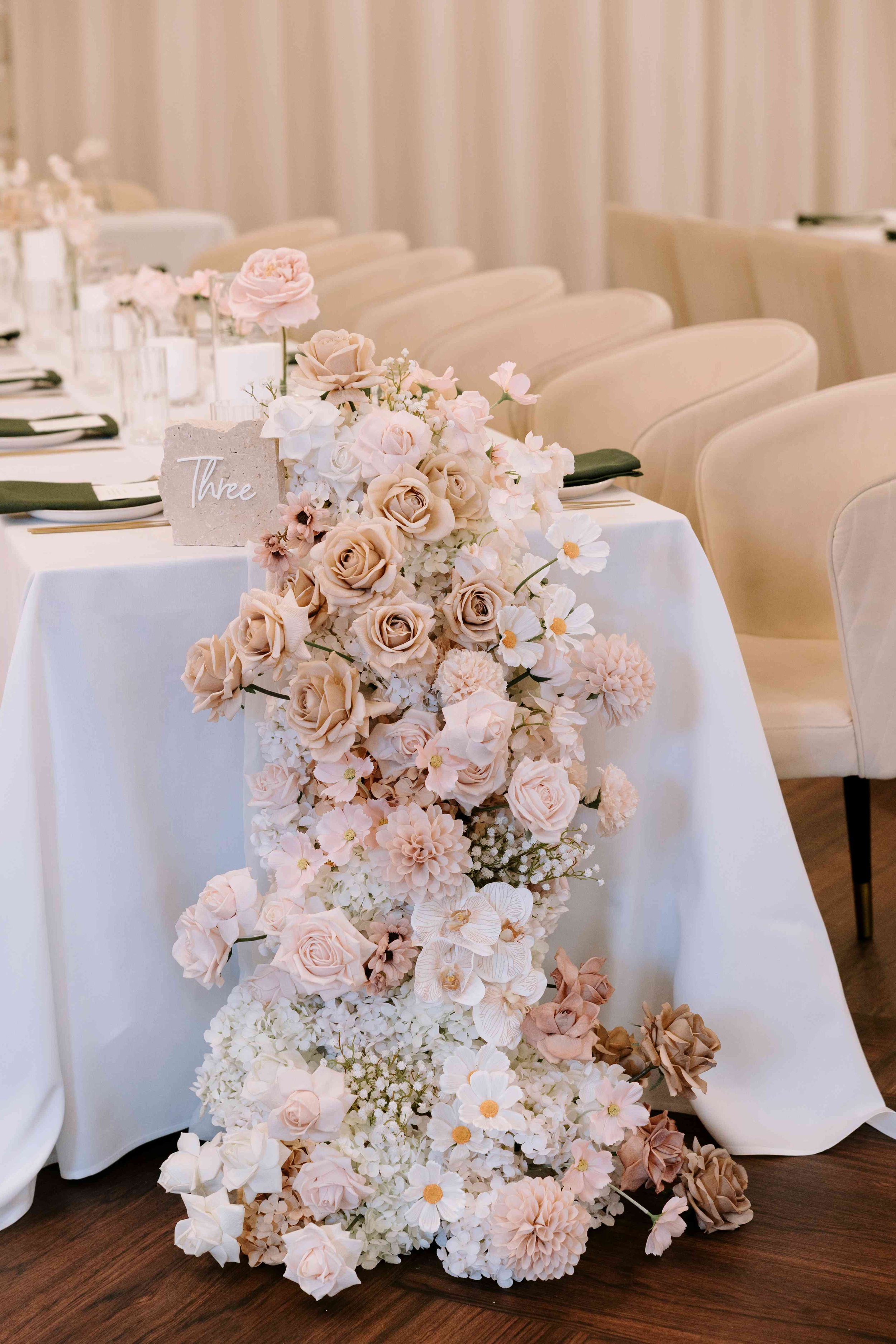 A cascading faux floral centerpiece at the end of the table with pink, white, and beige roses, dahlias, peonies, and orchids on a table at a formal event or wedding, with a table number card labeled 'Three'.