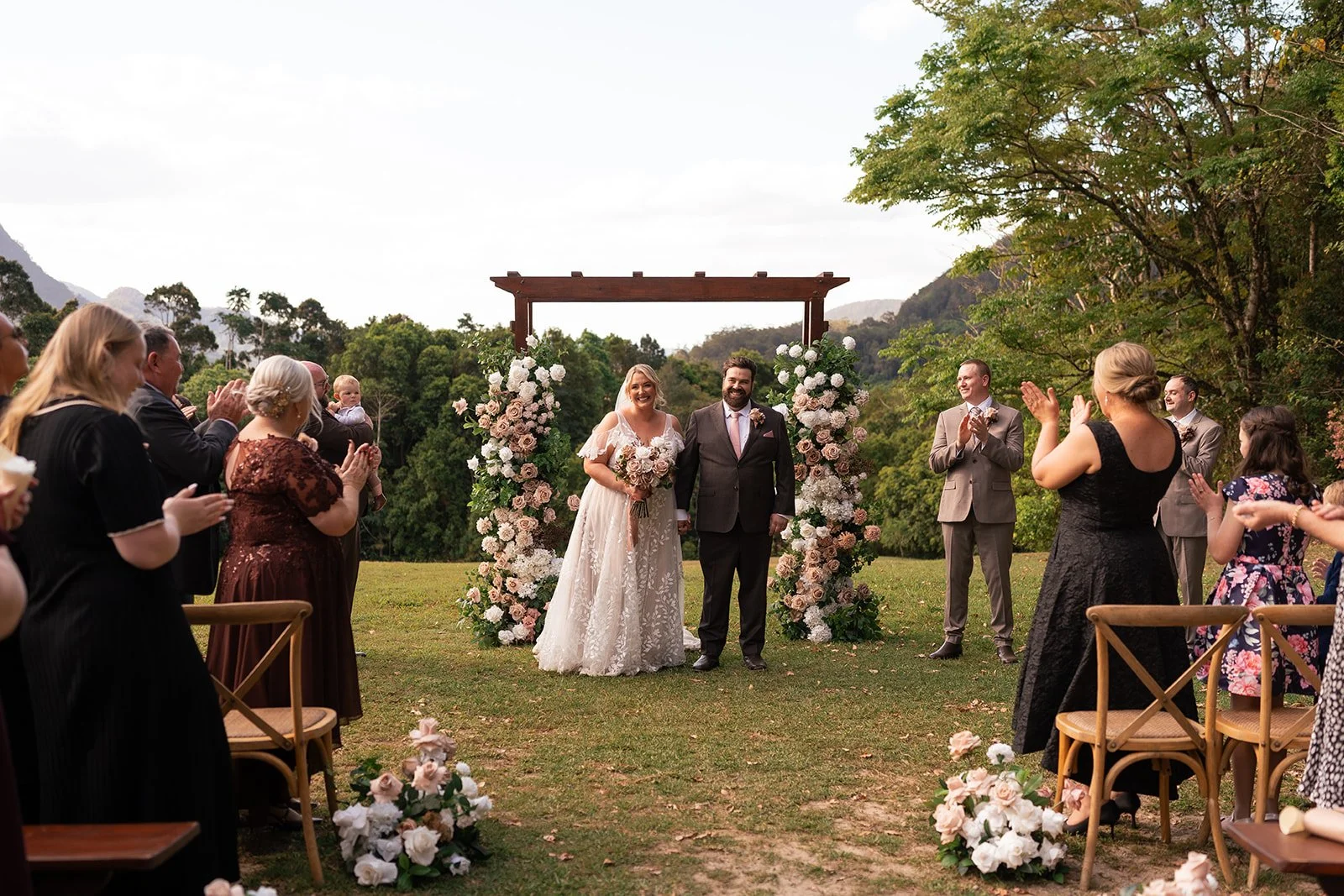 Bride and groom smiling under an artificial floral arch during their outdoor wedding ceremony, surrounded by guests clapping and celebrating in a scenic outdoor setting with trees and mountains in the background. Mocha toned flowers.