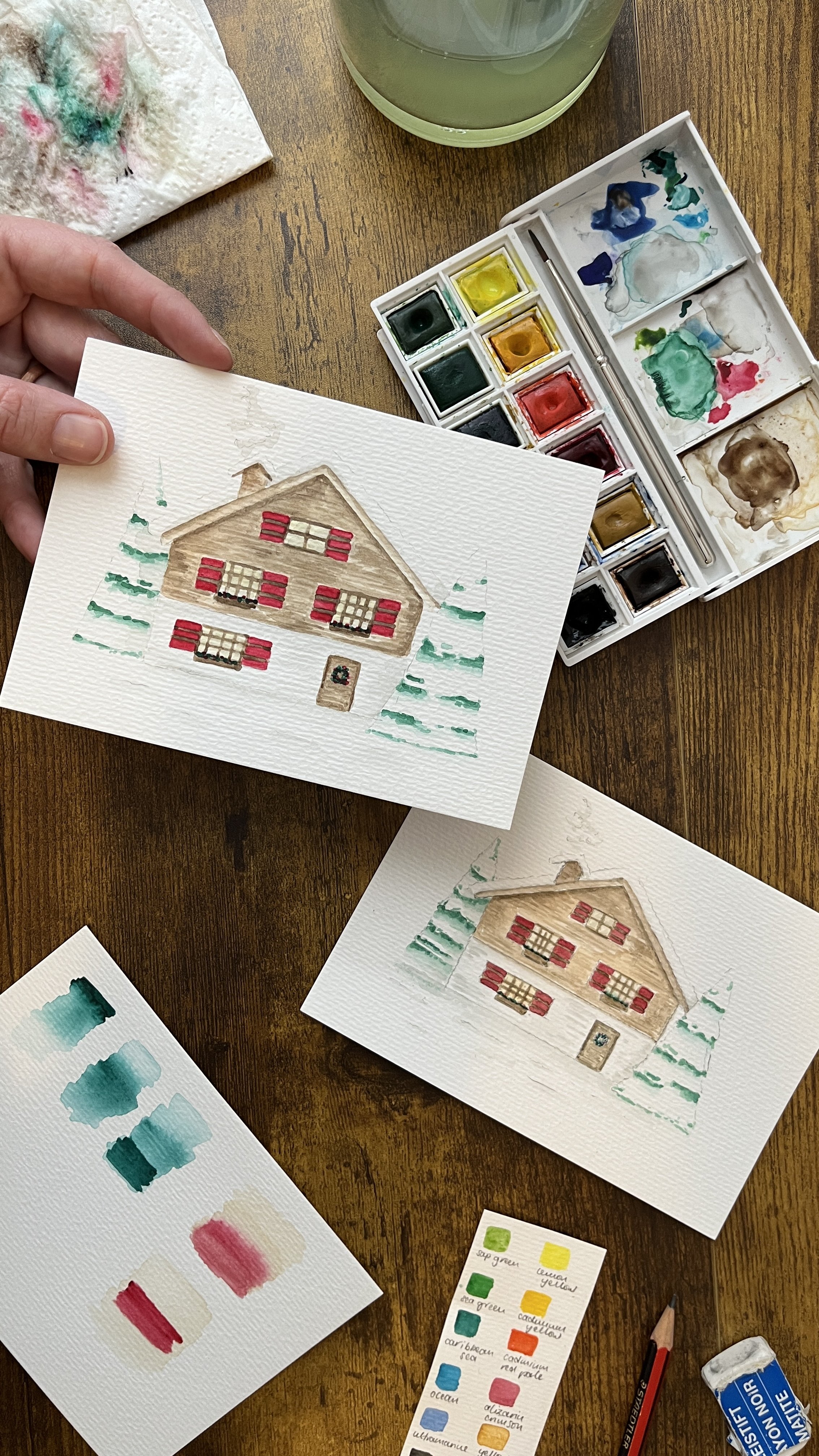 A photograph of two similar watercolour paintings by Victoria Salerno. They are both of a Swiss chalet surrounded by snow and a pine tree on either side. One painting is on the desk, next to a watercolour set, the other in one hand above the desk.