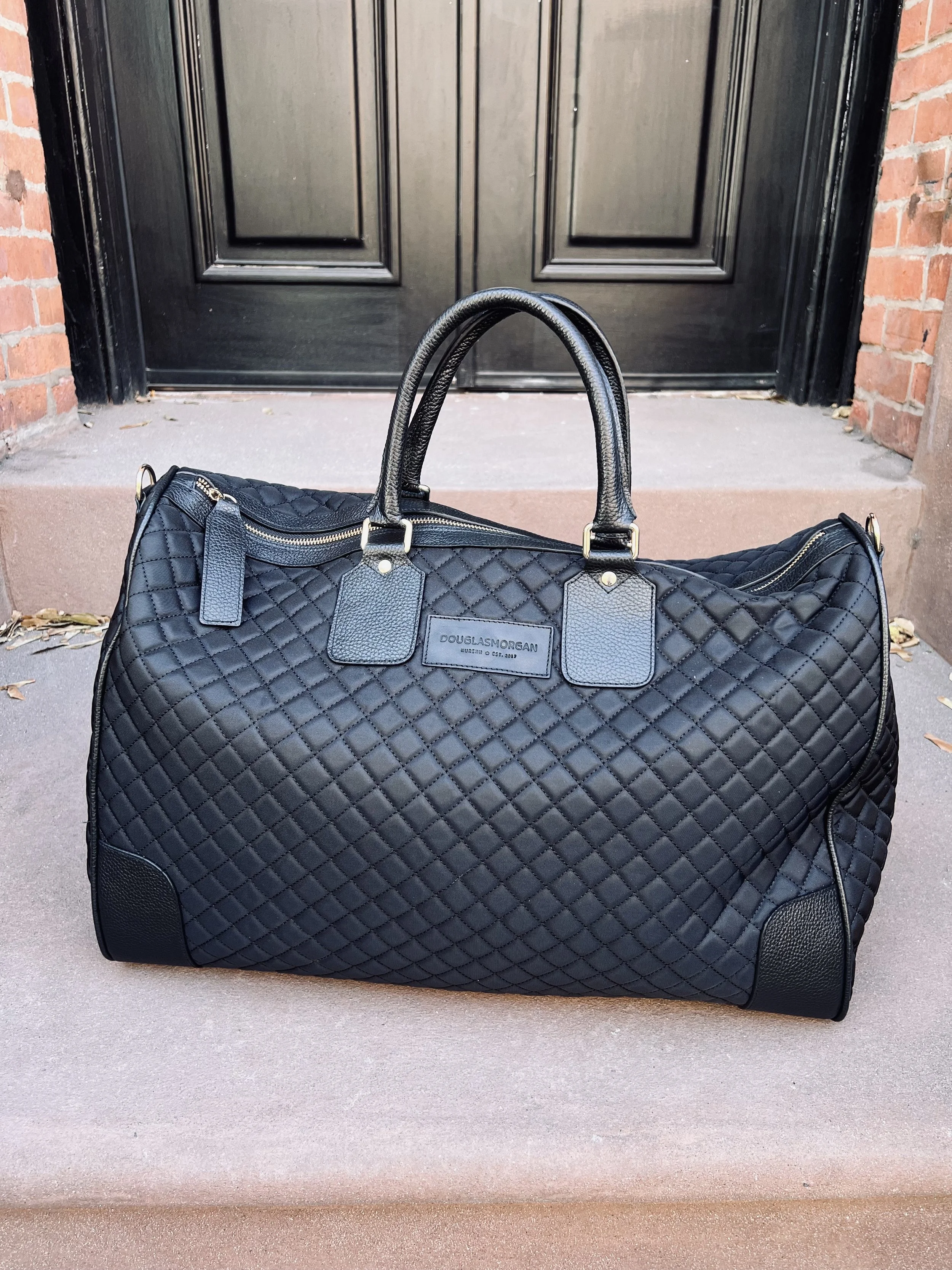 Black quilted leather duffle bag with gold zipper and handles, placed on a concrete step in front of a black door with brick walls on both sides.