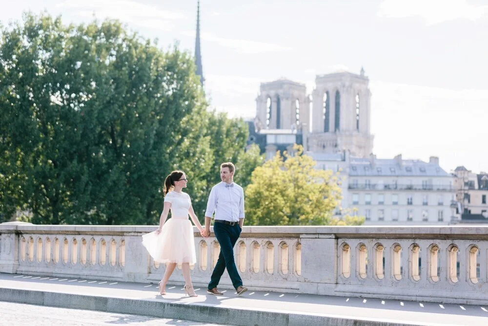 A couple holding hands walking on a bridge with a historic building in the background, trees, and a partly cloudy sky.