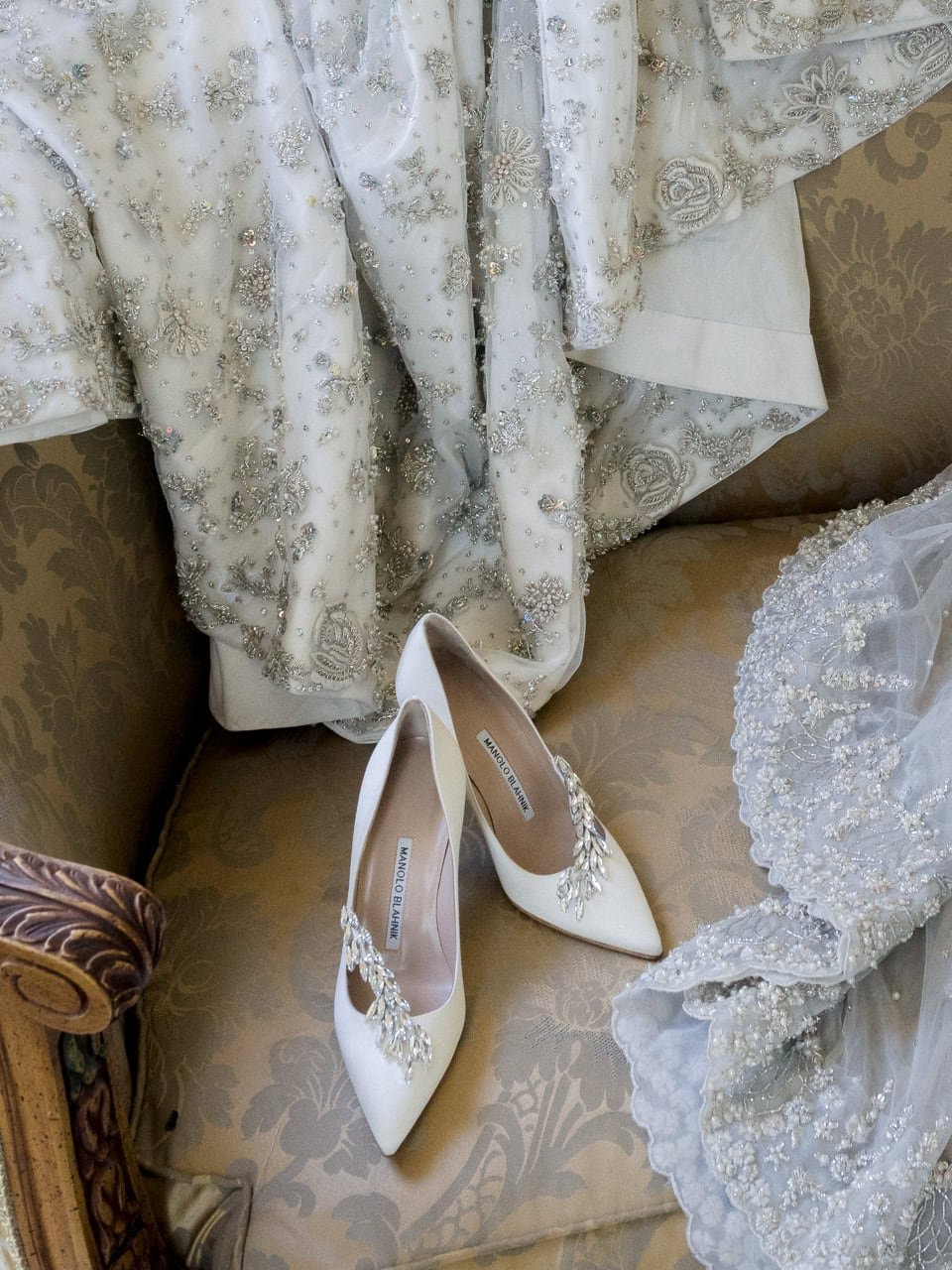 Embellished wedding dress with floral lace details and ornate high heels on a patterned chair.