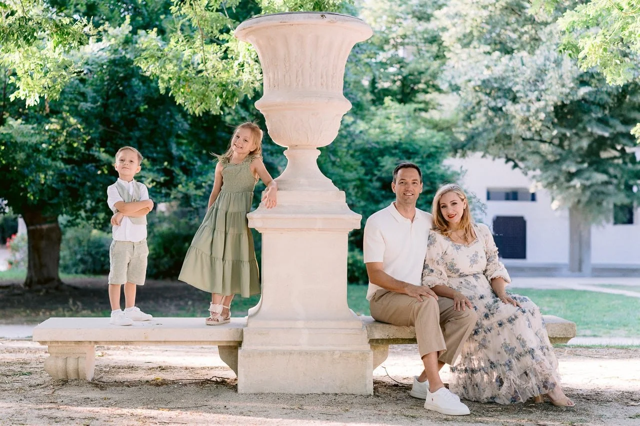 A family of four sitting on a park bench with trees in the background, smiling for the camera. The father and mother are sitting together, and two children are standing beside a large decorative stone urn.