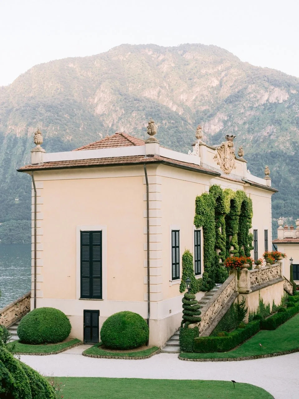 A grand villa with decorative roof ornaments, black window shutters, manicured bushes, and lush green grass, set against a mountain backdrop.