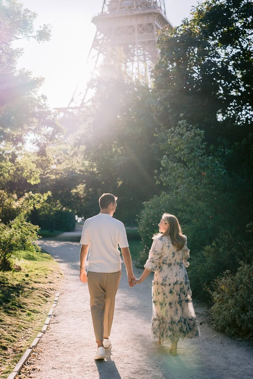 A couple holding hands walking down a pathway at sunset, with the Eiffel Tower partially visible through trees in the background.