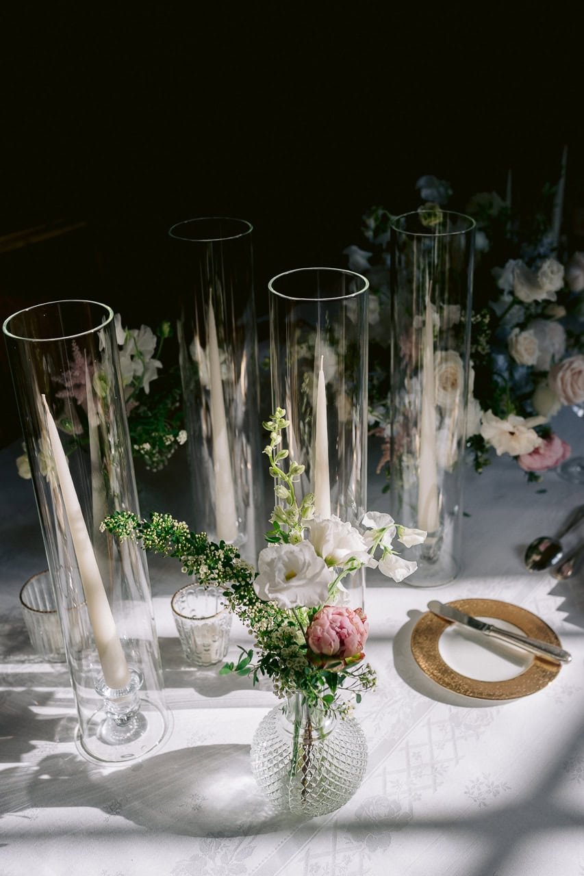 Elegant table setting with tall glass candle holders, white taper candles, a decorative glass vase with white and pink flowers, and a place setting with a gold-trimmed plate and cutlery.