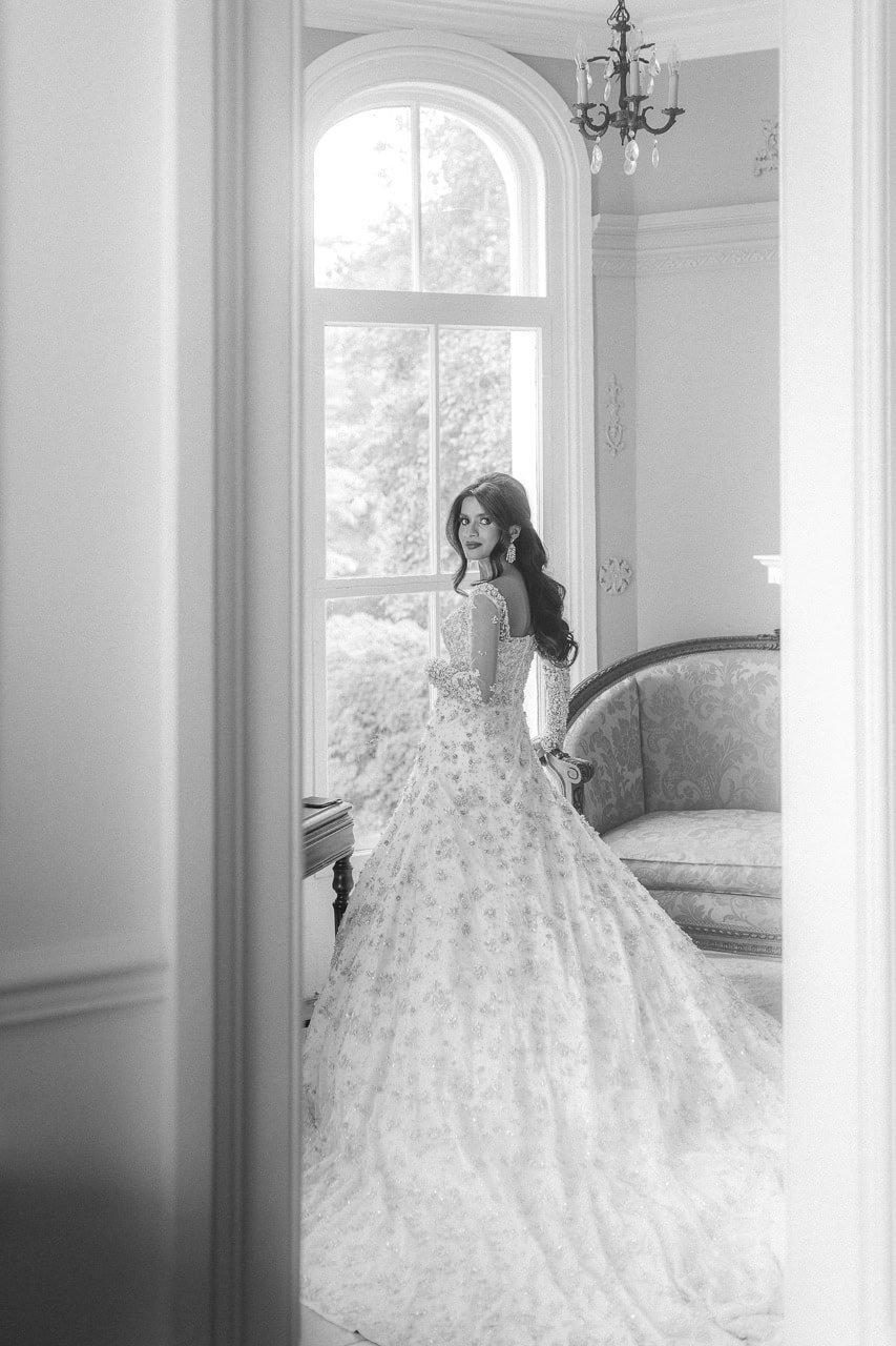 Black and white photo of a woman in a wedding dress standing by a large window, with ornate furniture and a chandelier in the background.