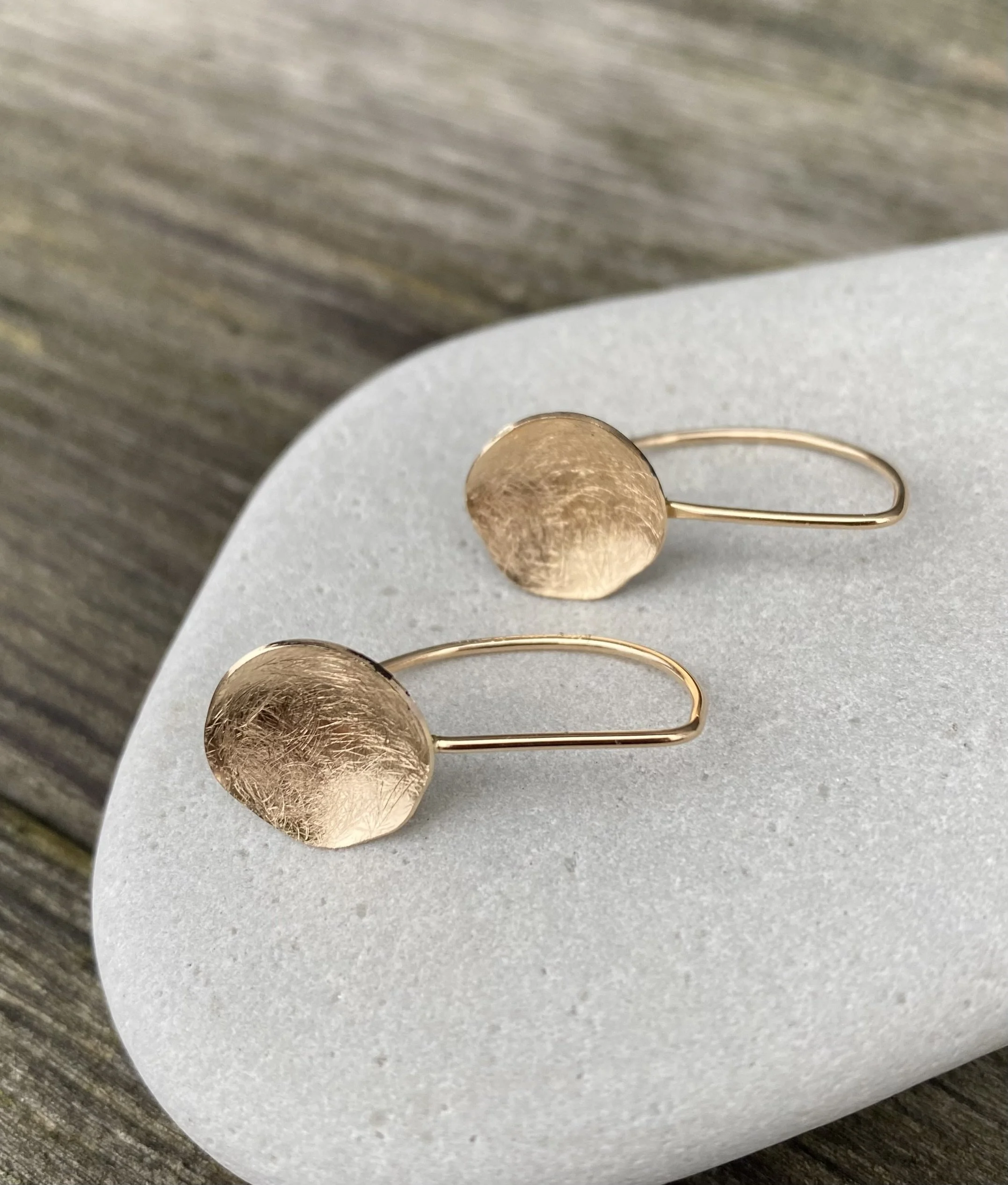 Ett par guldörhängen med texturerade oregelbundna runda konkava skivor, placerade på en ljusgrå yta.