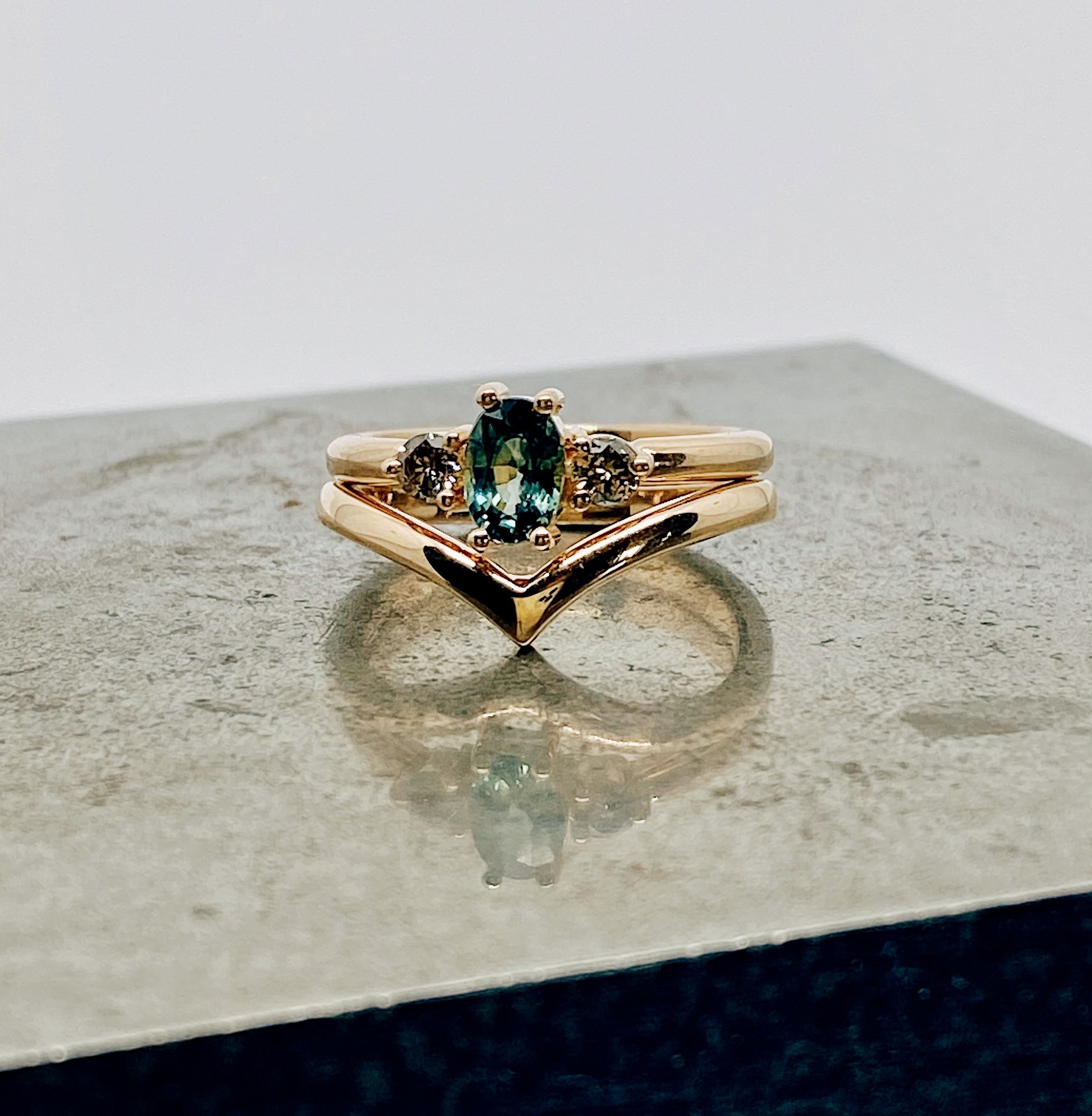 En guldring med en central oval teal-färgad safir flankerad av två små bruna diamanter, en undre ring med en V-formad design, wishbone-ring, placerade på en texturerad yta.