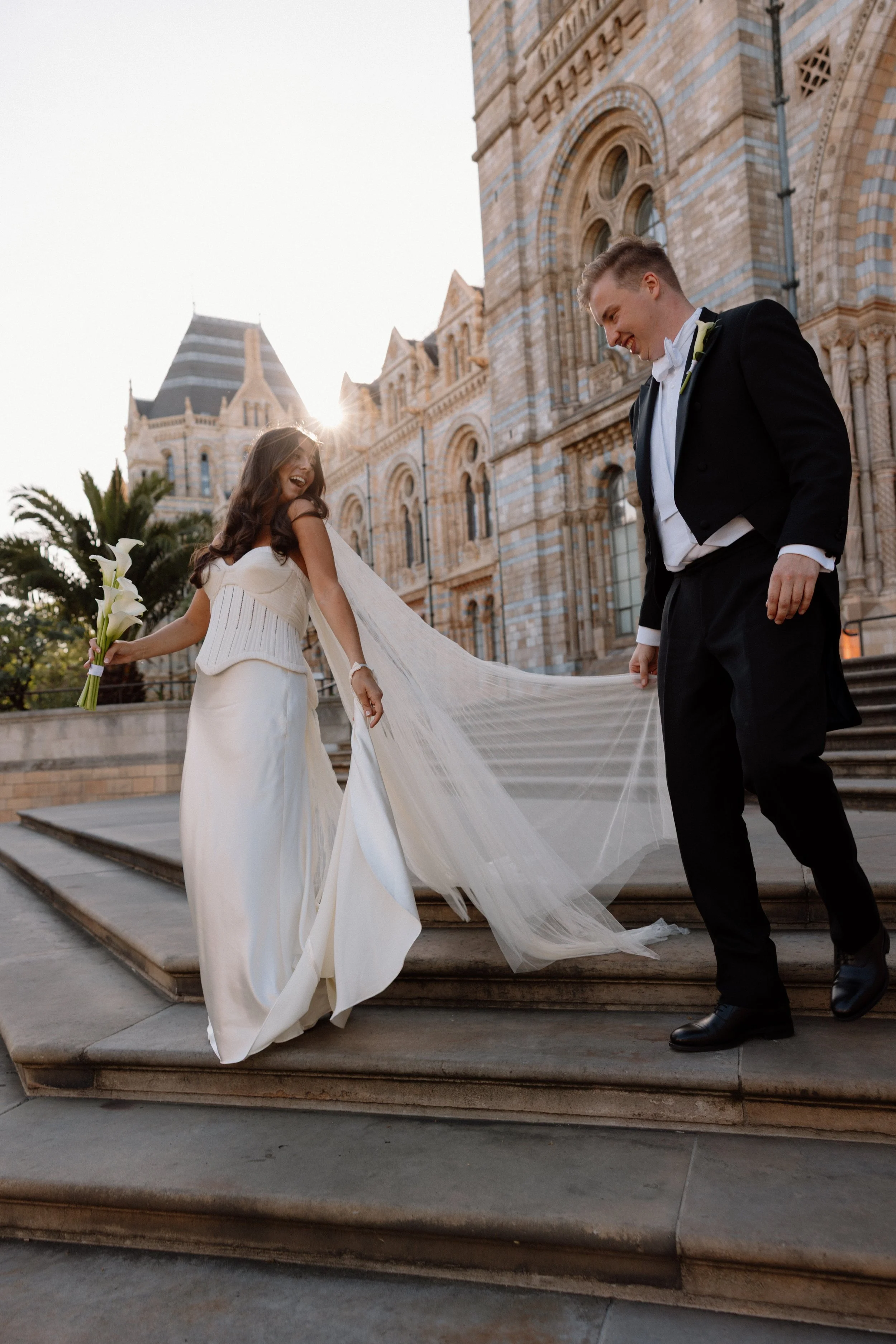 A bride in a white wedding gown holding a bouquet of calla lilies, and a groom in a black tuxedo, standing on steps outside a historic building with ornate architecture, during sunset.