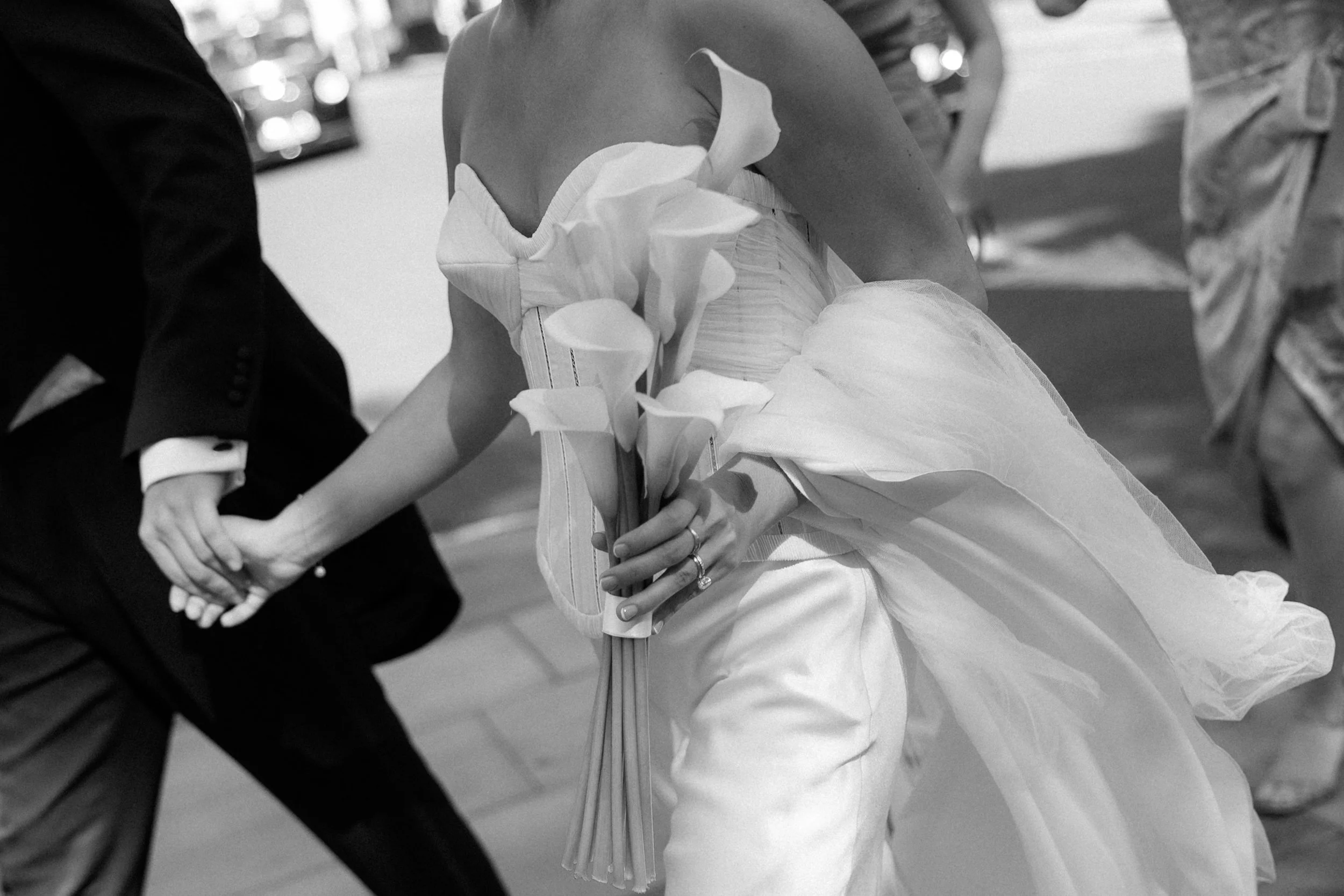 A black-and-white photo of a bride and groom holding hands, with the bride wearing a strapless wedding dress and holding a bouquet of calla lilies, at a wedding ceremony.