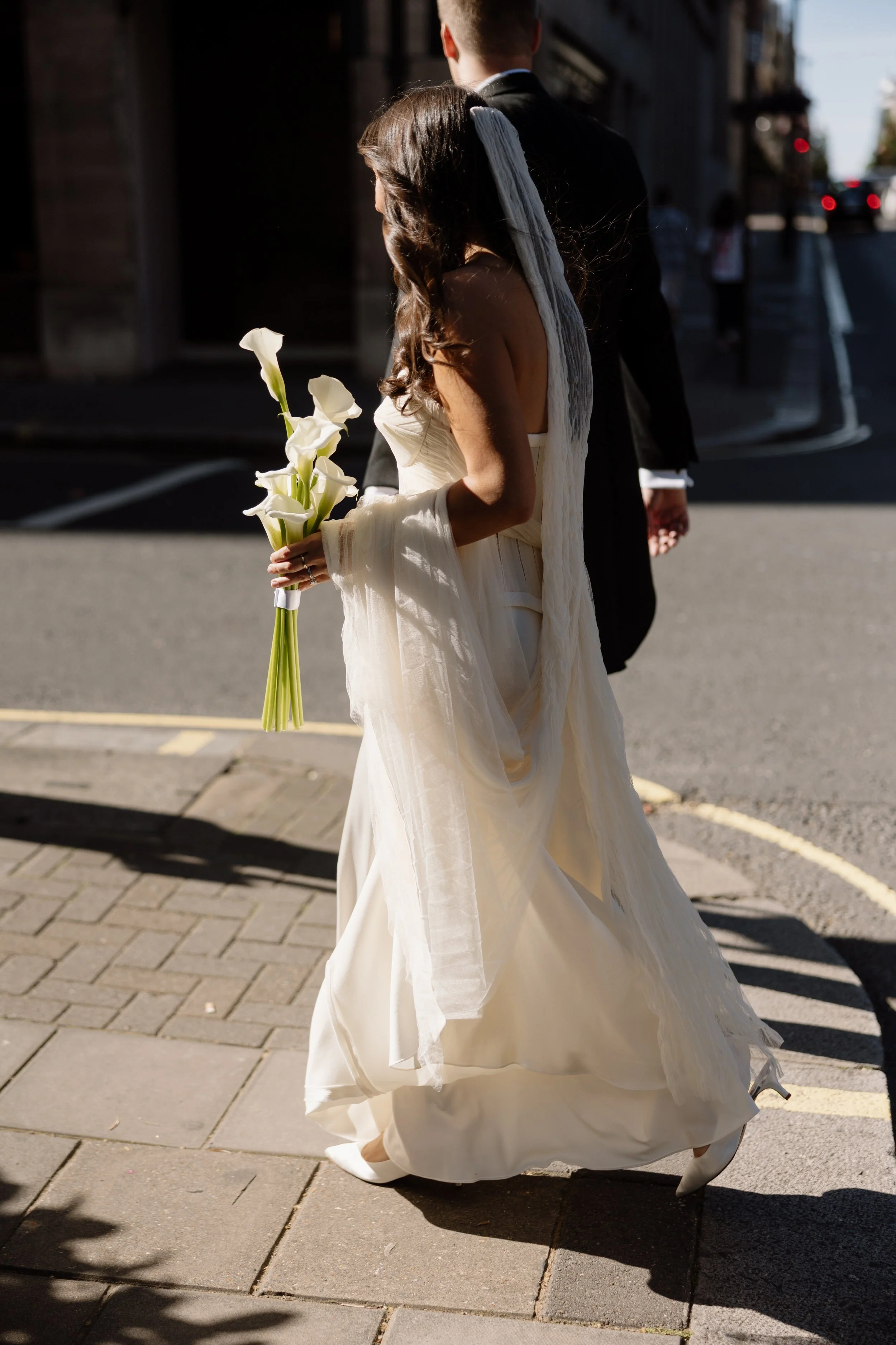 A bride in a white dress holding a bouquet of white calla lilies walks on a city sidewalk. She is walking with a groom in a dark suit. The photo captures the back view of the bride and groom, with sunlight casting shadows on the ground.