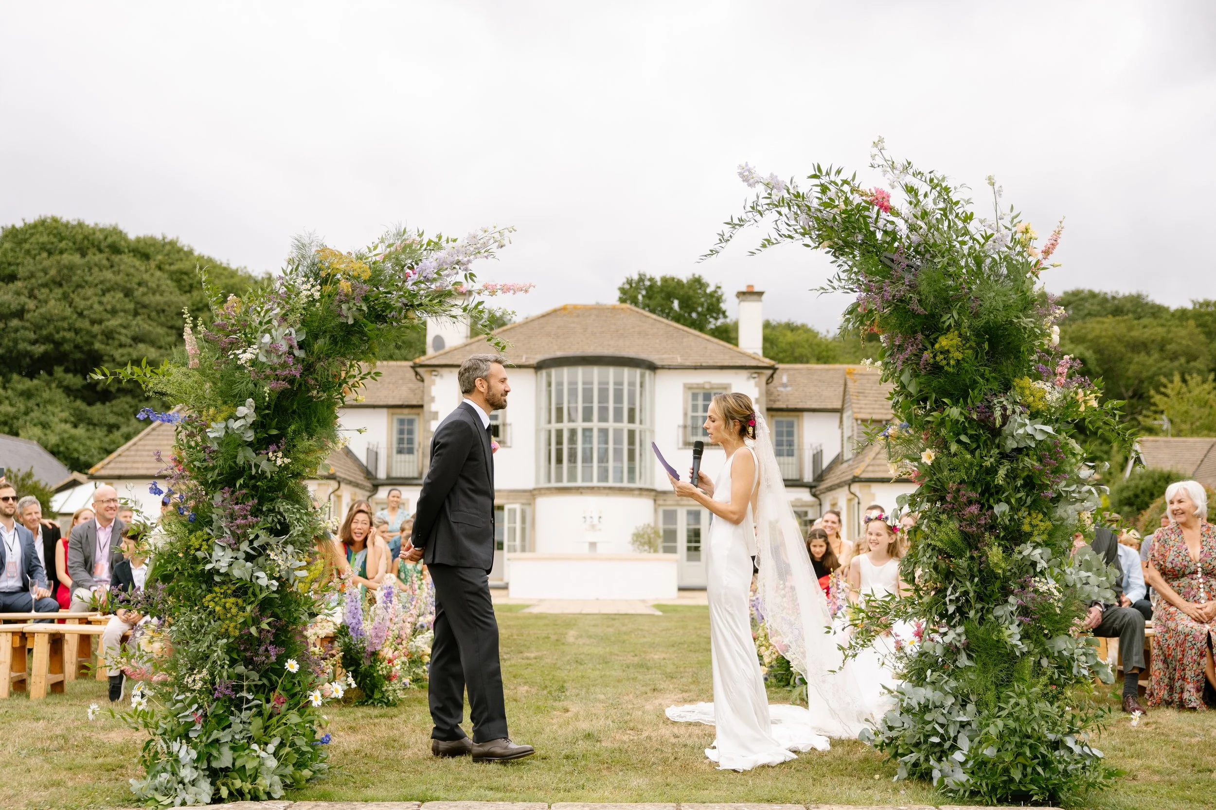 A bride and groom exchanging vows during an outdoor wedding ceremony, with a floral arch behind them and guests seated on either side.