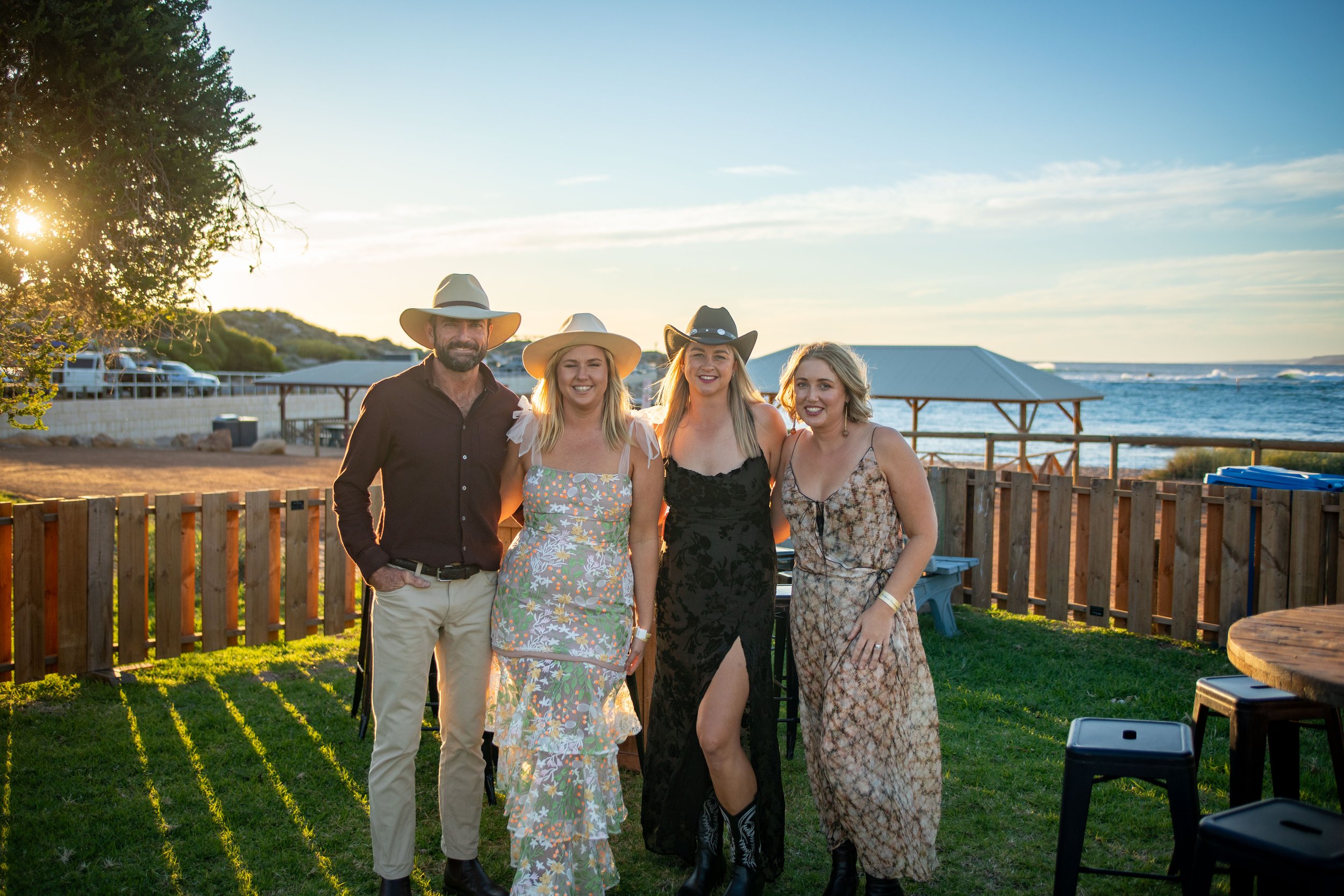 Four people posing outdoors near a beach with a wooden fence, trees, and a gazebo in the background, during sunset or late afternoon. Kalbarri gala