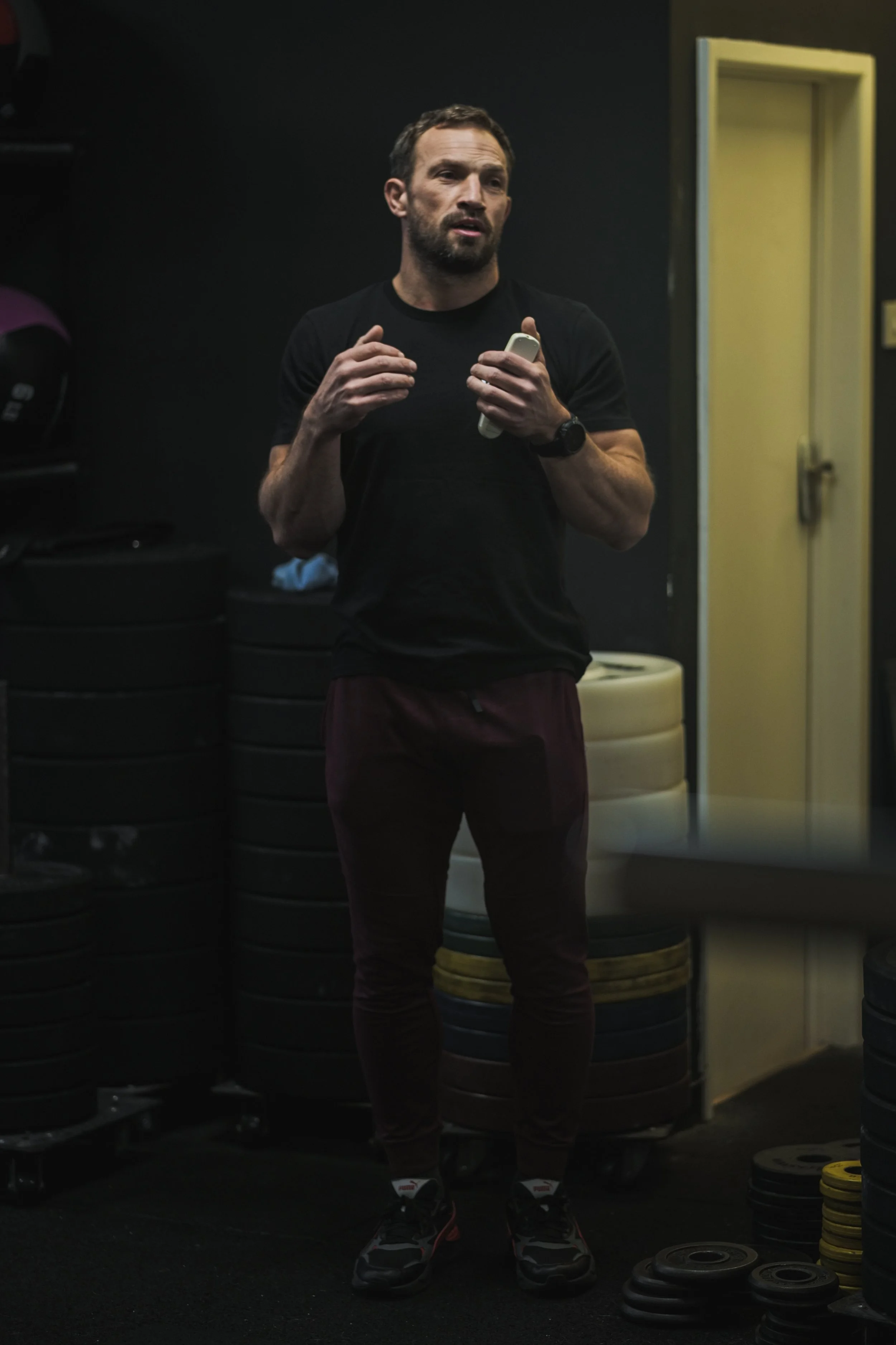 A man in athletic wear, holding a phone, standing in a gym with weight plates stacked in the background.