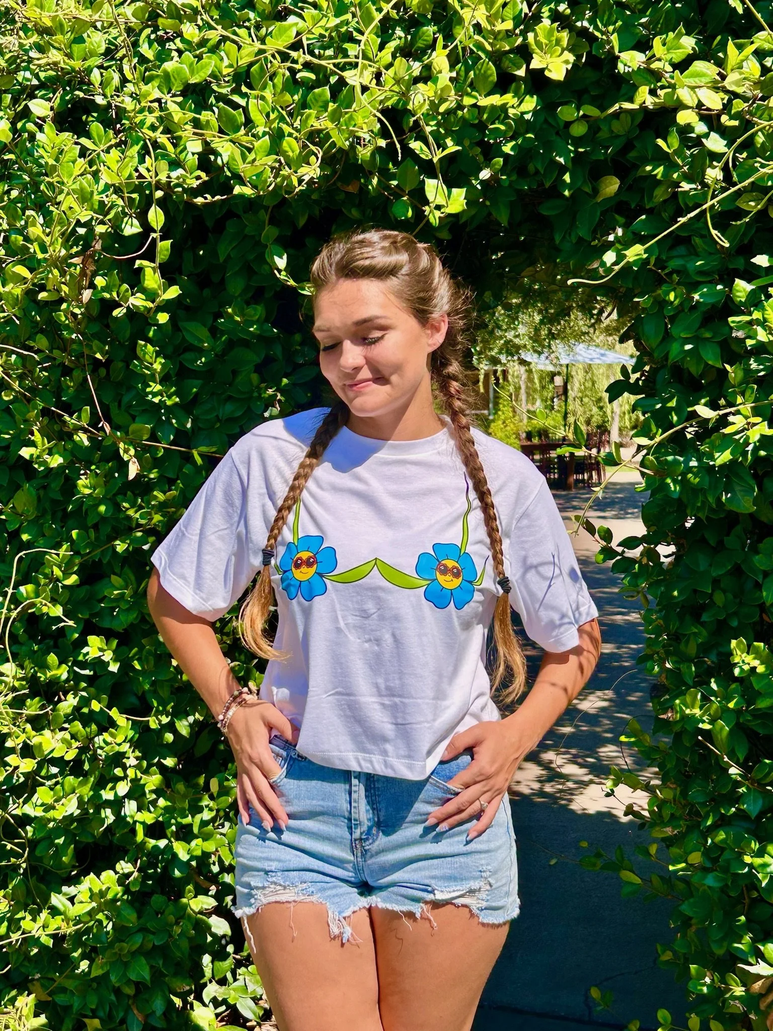 young lady in braids wearing a white shirt with a flower design in the shape of a bra. she is standing under a living plant archway