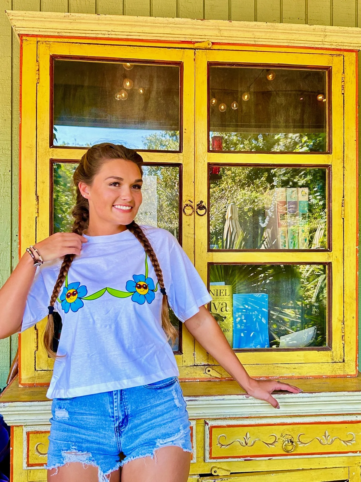 young lady with braids wearing a white t shirt with a blue flower bra design. standing in front of a yellow bookcase.