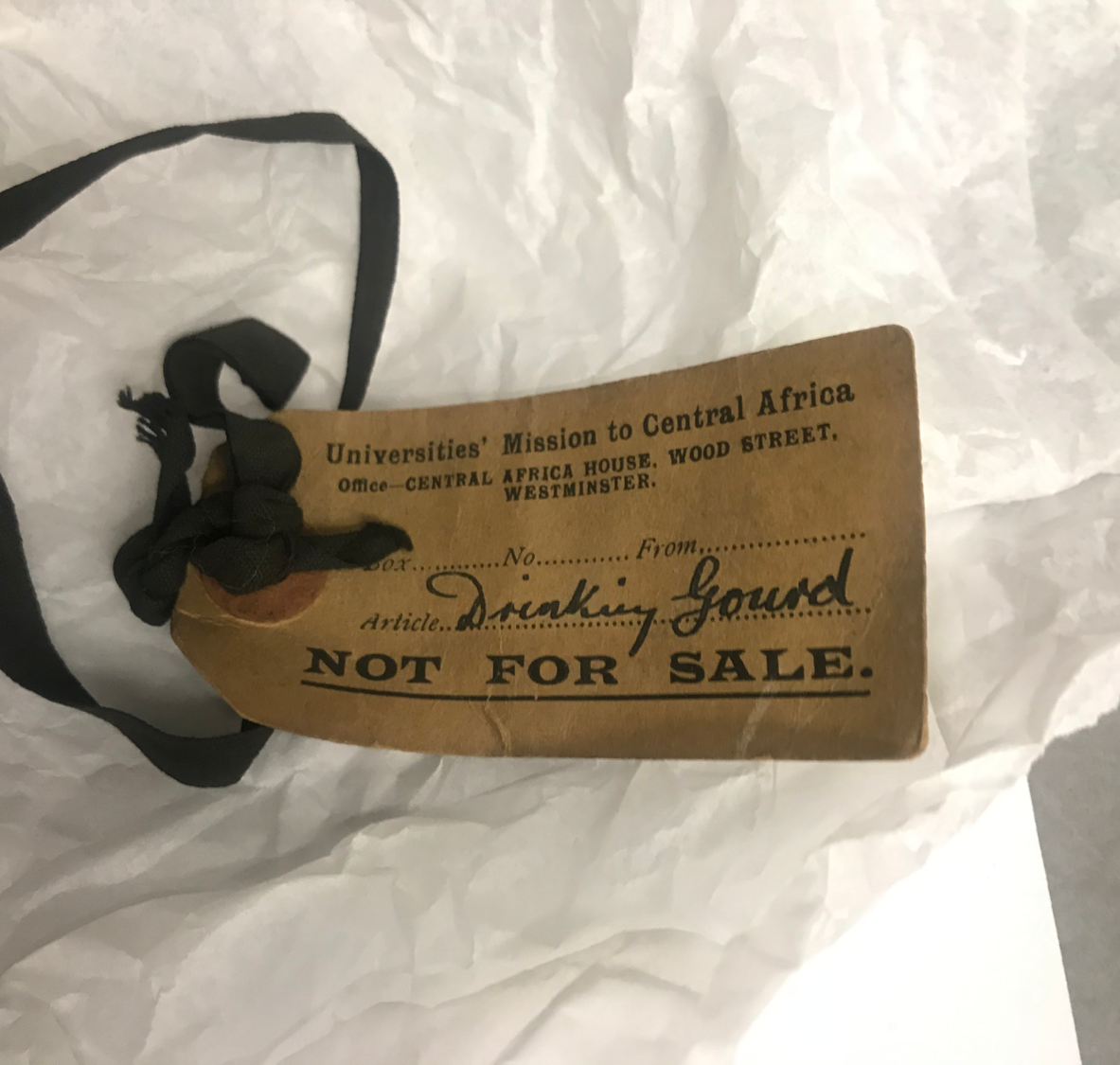  A label on aged paper with a leather thong tied through one end. The label reads: “Universities’ Mission to Central Africa Office- Central Africa House, Wood Street, West Minster Box....No.....From....Article Drinking Gourd NOT FOR SALE”. 