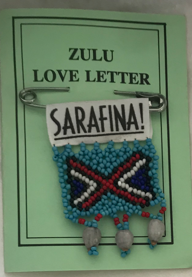  A light green paper booklet with the title in capitals: 'Zulu Love Letter'. A safety pin has been pierced through it. Attached to the safety pin is a small Zulu style beaded badge made up of light blue beads with two white-outlined, black triangles 