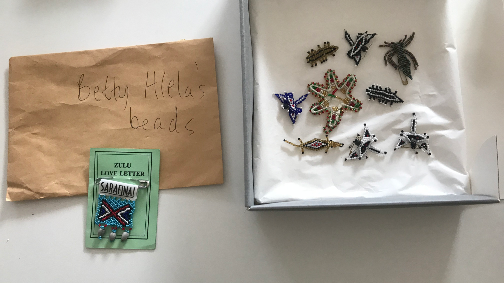  On the left is a brown envelope which reads 'Betty Hlela's beads' in handwritten biro pen. The Zulu love letter card with the safety pinned Zulu beaded badge is placed on top of it. On the right is a box of nine beaded badges in the shape of dragonf