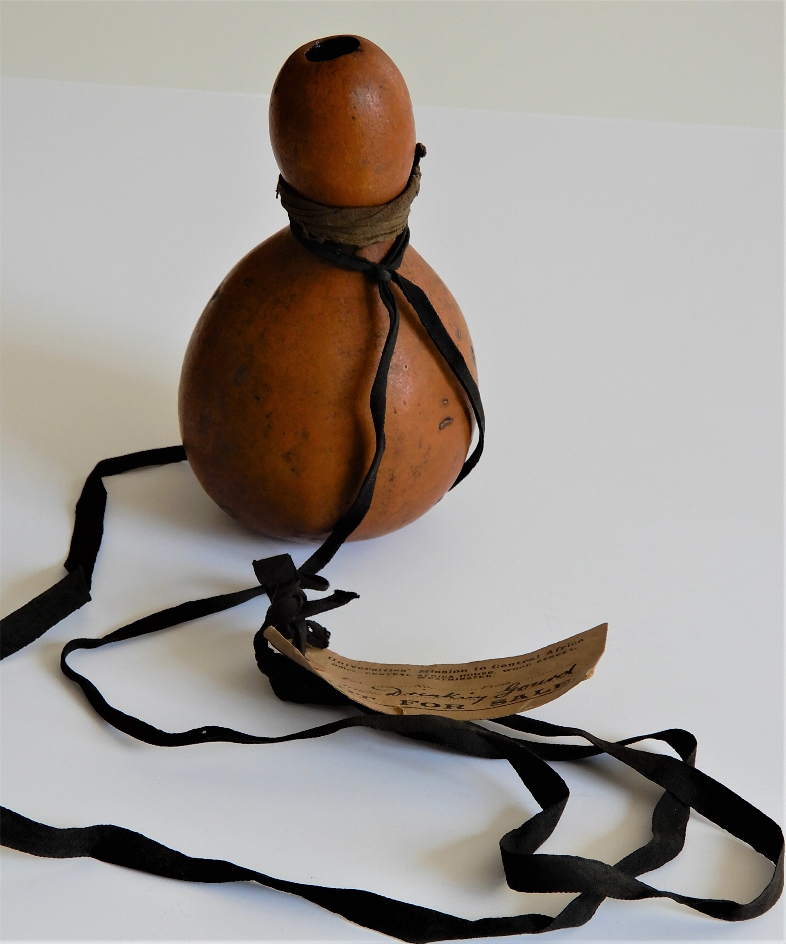  A medium brown gourd which has one large bulbous shape at the bottom and a more elongated slimmer bulbous shape at the top. At the narrowest point there is a leather strip wrapped around it. The gourd sits on white protective wrapping paper. 