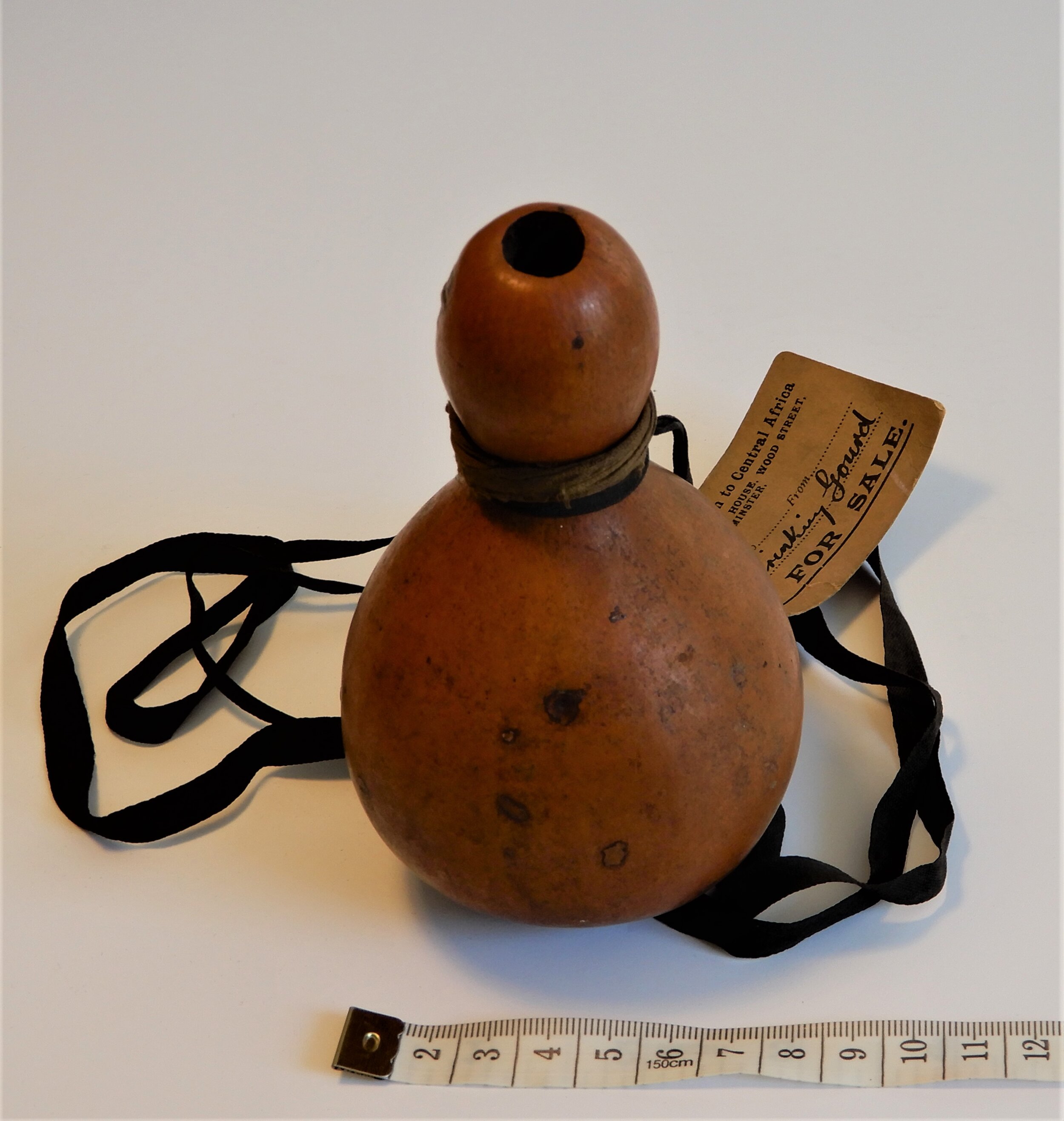  A medium brown gourd which has one large bulbous shape at the bottom and a more elongated slimmer bulbous shape at the top. It. is viewed from the top so you can see the hole in the top.   Closer to the viewer is an old fashioned measuring tape. 