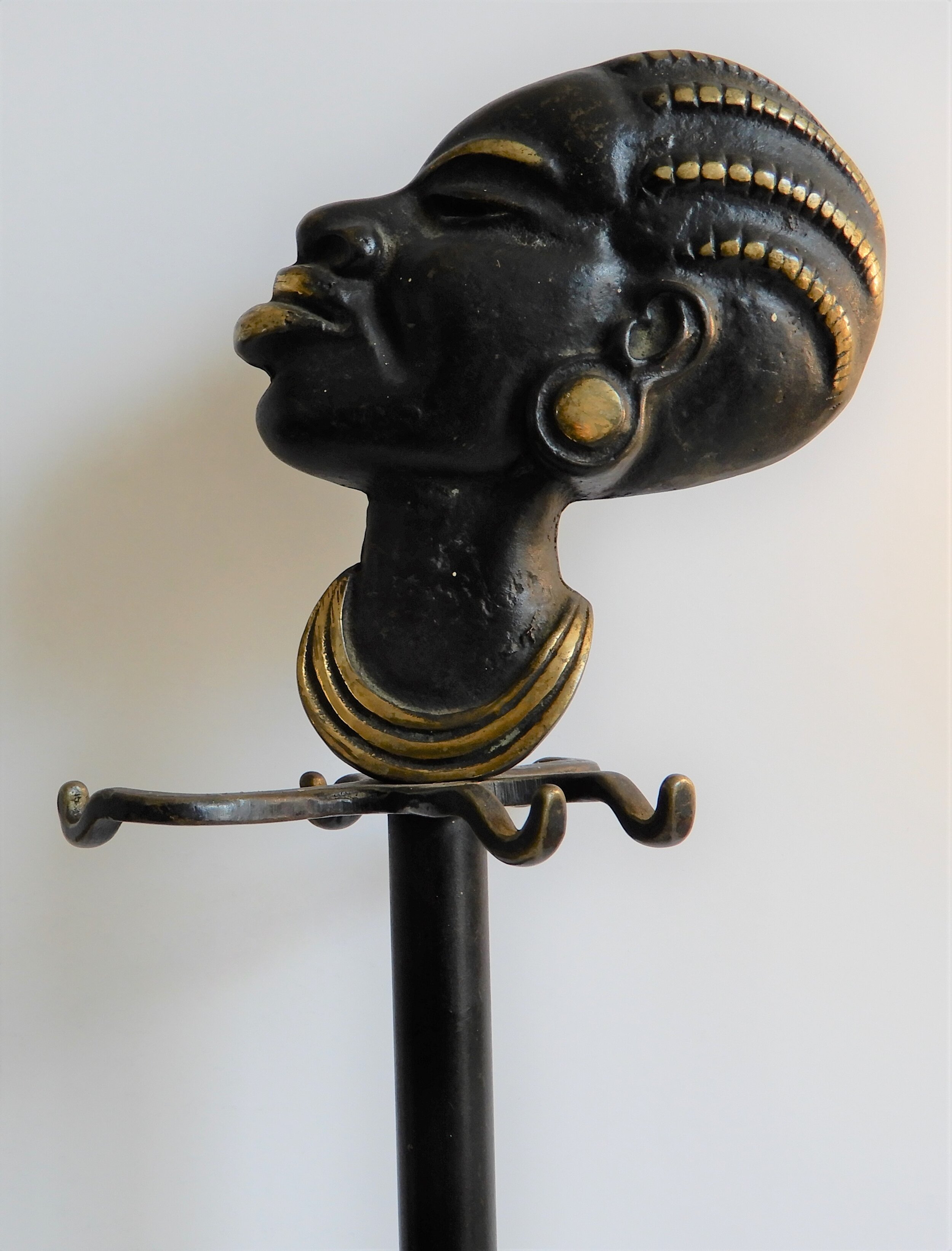  A black fire iron set holder the top of the stand are four hooks, three at the front and one at the back. At the top there is a sculpture of a Black woman in profile. Her fours braids, eyebrows, lips and necklace are painted gold. Her eyes are close