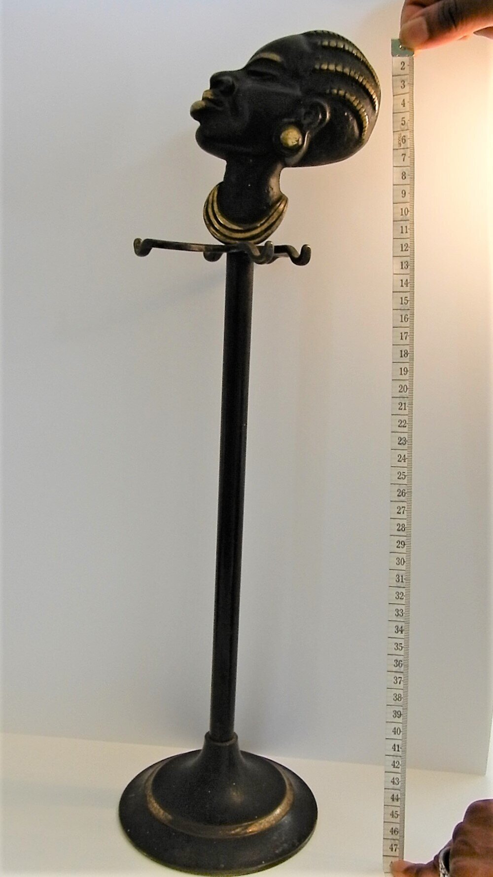  A black fire iron set holder showing the tall stand. Towards the top of the stand are four hooks, arranged radially around the stand. At the top there is a sculpture of a Black woman in profile. Her fours braids, eyebrows, lips and necklace are pain