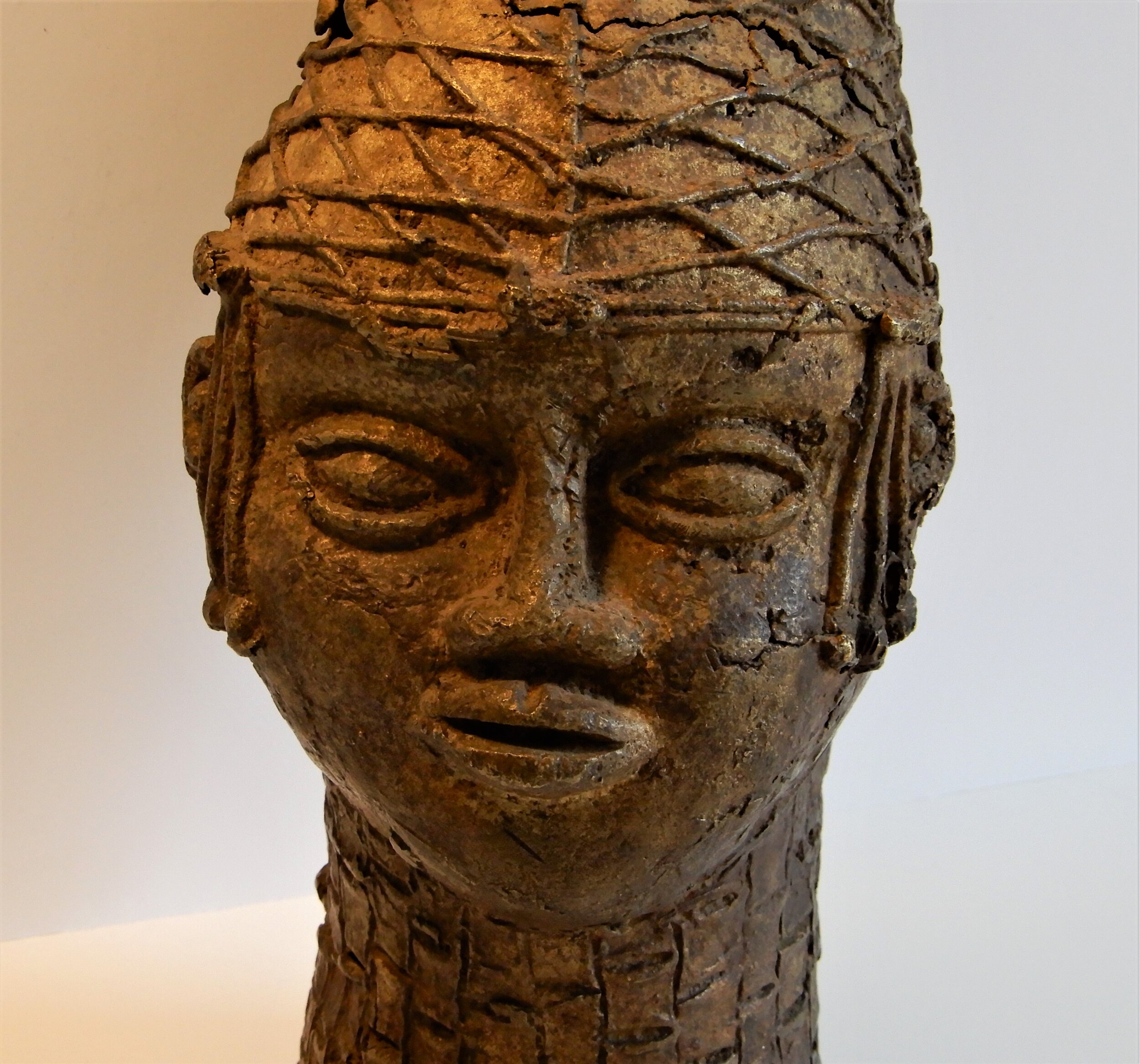  A close up of a bronze bust of a feminine face. Her hair is styled as an elegant cone slightly curved forward. Around the cone are criss crossed strips of bronze. 