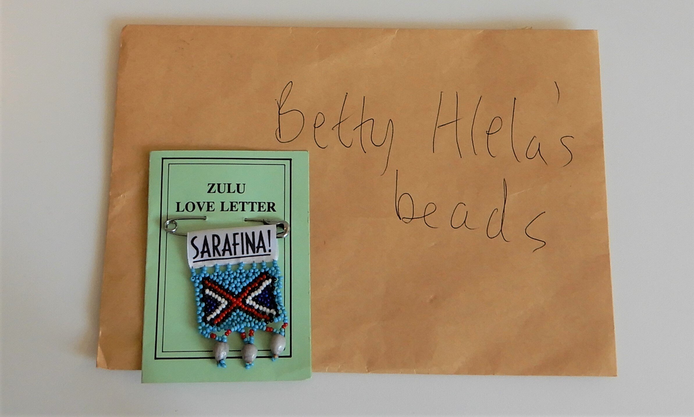  A brown envelope which reads 'Betty Hlela's beads' in handwritten biro pen. The Zulu love letter card with the safety pinned Zulu beaded badge is placed on top of it on the left. 