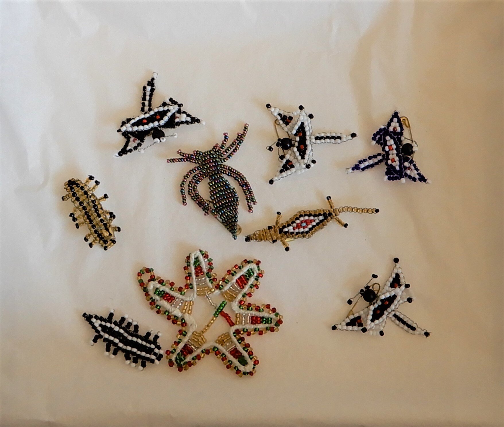  A display of nine beaded badges in the shape of dragonflies, caterpillars and crocodiles. 